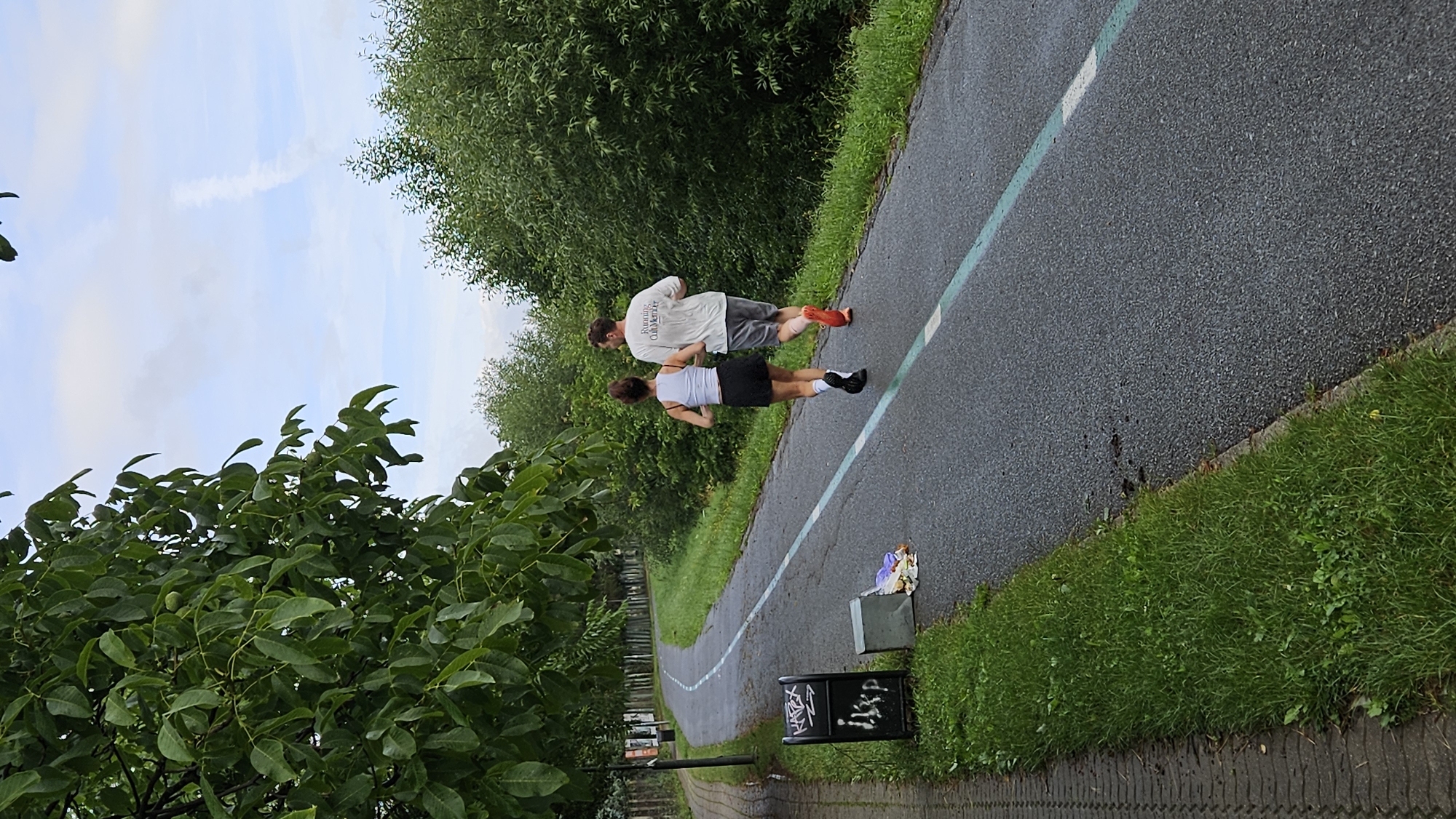 Two individuals, a man and a woman, are jogging on a paved path lined with a teal stripe. The man, wearing a light-colored t-shirt with text that appears to read "Running Club Kvantum" and grey shorts, is ahead of the woman. The woman is wearing a white tank top and a dark skirt. They are moving away from the camera. To the left of the path, there is a trash can with graffiti, and next to it, a small grey concrete object with some discarded trash, including plastic bags and wrappers. The path is bordered by a grassy area and a sidewalk made of interlocking pavers. Beyond the path and grass, there are lush green trees and foliage, suggesting a park or a greenway. The sky above is partly cloudy with a thin contrail visible. The scene appears to be taking place in an outdoor urban setting. The location is identified as the vicinity of Lúčna Street in Prešov, Slovakia, based on the presence of a similar running path and signage visible in the background which matches descriptions and visual references of this area.