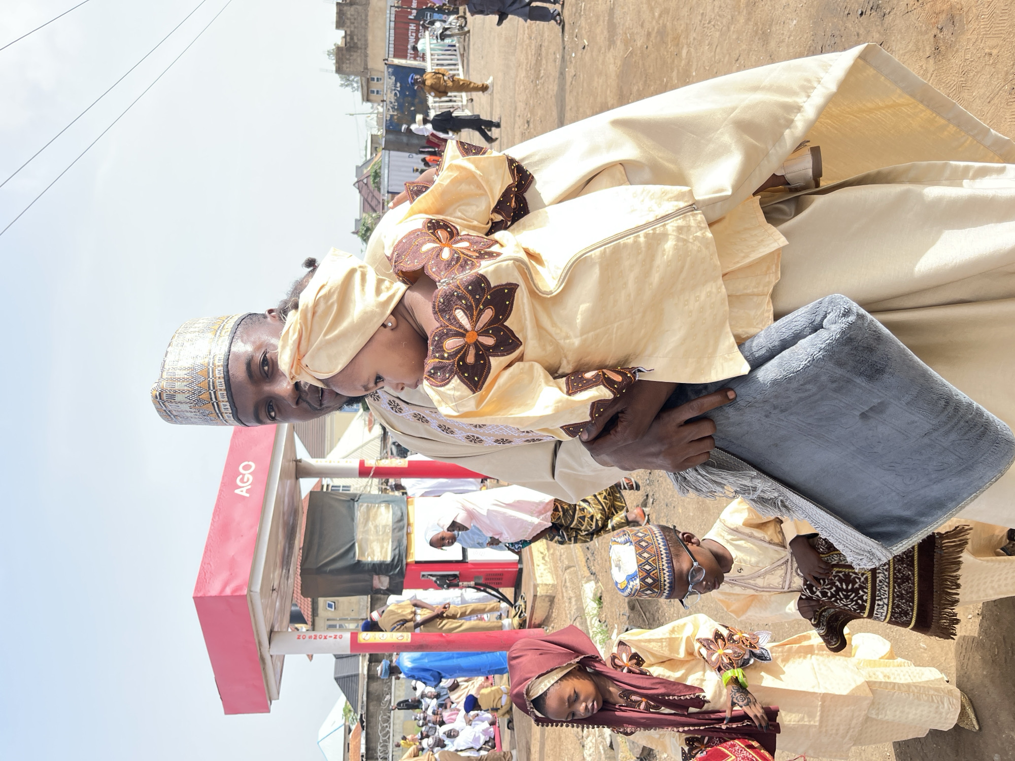 The image shows several individuals in Jos, Nigeria, possibly attending an event or gathering. The style of dress suggests a cultural or religious occasion.
Several individuals wear traditional attire. Some individuals stand near what appears to be an AGOS gas station. Additional people are blurred in the background. The weather is sunny and it seems to be daytime.