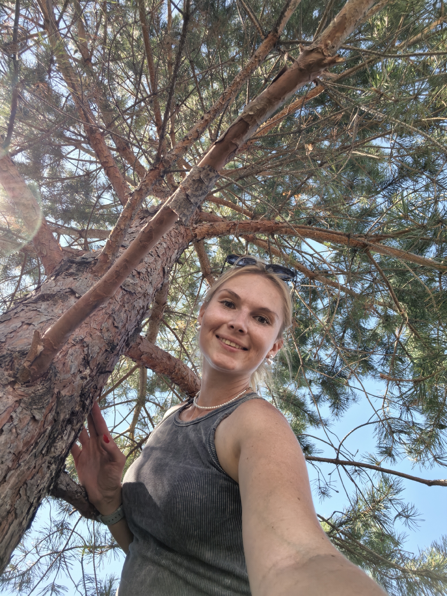 A young blonde woman is posing for a selfie outdoors, leaning against a large tree. She is wearing a grey ribbed tank top and a pearl necklace. Sunglasses are perched on her head. Her left hand is on the tree trunk, with red nail polish visible. The background consists of the tree's branches and pine needles, with a clear blue sky. The image appears to be taken on a sunny day. The location is not specified in the image itself, and therefore cannot be determined to be Sterlitamak, Russia.