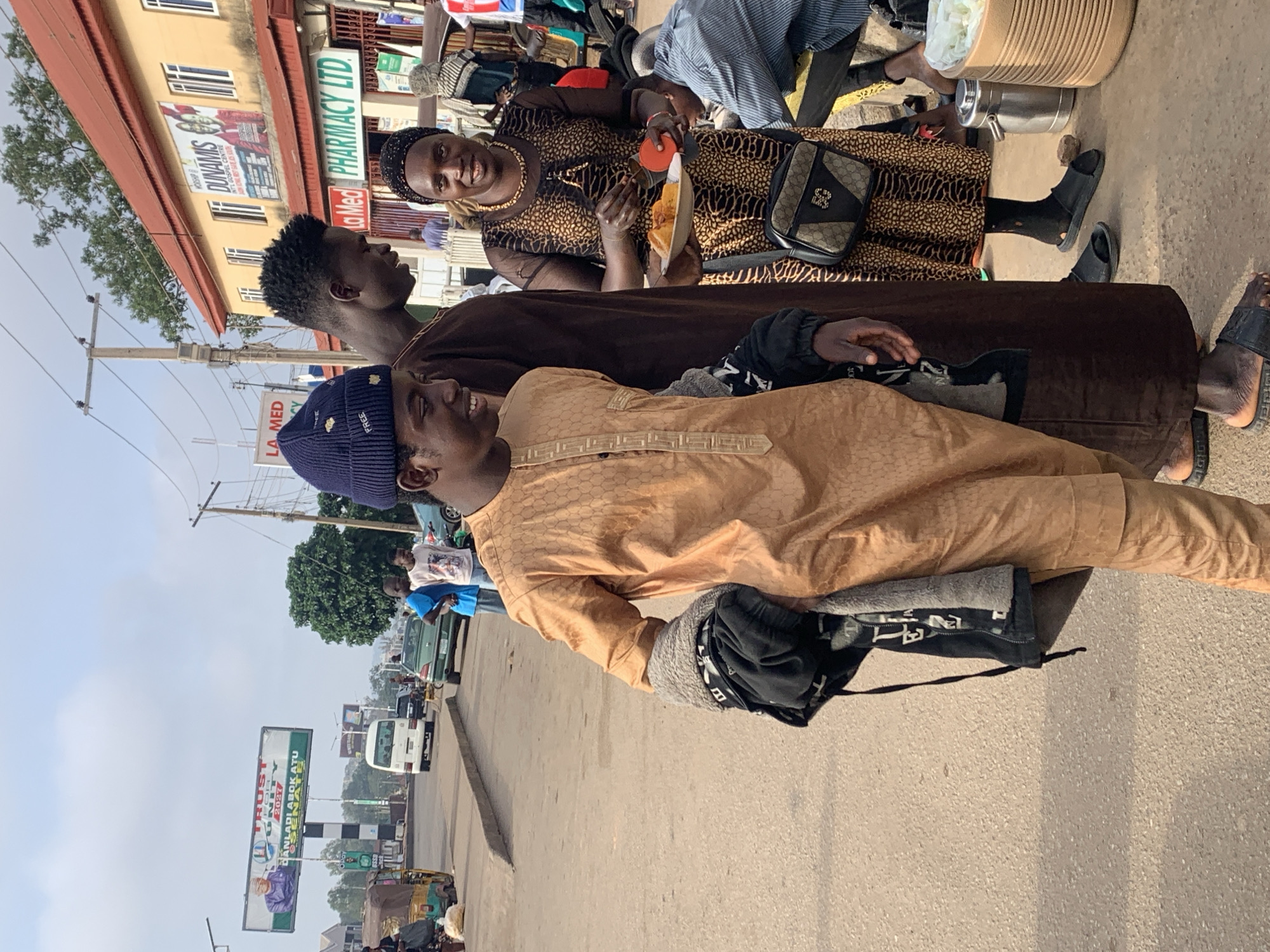 The image captures a street scene in Jos, Nigeria. In the foreground, a young man wearing a tan, patterned traditional outfit and a blue beanie is walking. He is carrying a black jacket over his left arm. To his right, another young man in a brown traditional outfit stands behind him. A woman wearing a leopard-print dress and a headscarf is to the right of the young men, holding a plate of food. She is looking towards the viewer and smiling. In the background, several vehicles, including a white minibus and a green car, are visible on the road. Buildings line the street, one of which has signage for "DUNAMIS" and "LA MED PHARMACY LTD". The overall environment appears to be a busy market or commercial area.