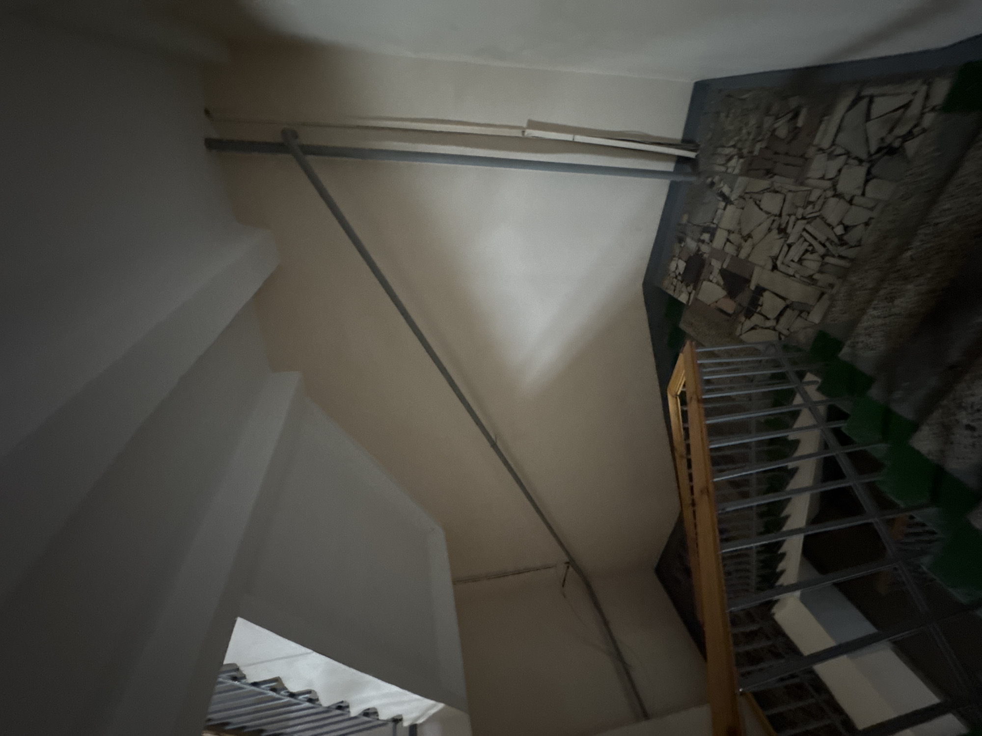 The image shows an indoor view of a stairwell with a railing. The stairs are partially visible and appear to be made of concrete with green painted risers. The railing is made of metal with a wooden handrail. The walls are painted a light color. There are pipes running along the wall. The floor at the bottom of the stairs is tiled with irregular shaped stones. There are no people visible in the image. The location is in the city of Unknown, Unknown.