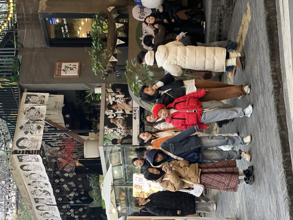 This image depicts a pedestrian street scene within a commercial area in Liangjiang, China. The environment consists of multi-level buildings constructed primarily of concrete or stone, featuring black metal railings on upper levels and various storefronts.
In the foreground and midground, approximately fifteen East Asian individuals are present, predominantly adults with at least one child. They are dressed in cool-weather attire, including various puffer jackets, long coats, hoodies, and a knitted hat.
Specific individuals observed:
* A young person in a bright red puffer jacket, gray sweatpants, and a red knitted hat is positioned centrally, facing slightly right.
* To their left, a young female in a tan puffer jacket and a red plaid skirt is looking down at a white smartphone. A white shoulder bag hangs from her right shoulder.
* Next to her, another young person in a blue denim jacket and dark blue hoodie is walking, carrying a light-colored cross-body bag.
* Further back, a woman in a long white puffer coat stands with her back partially to the viewer, while other individuals are grouped near storefronts, some appearing to converse or observe their surroundings. One person is seated inside a visible establishment.
* To the far left, a woman in a black puffer coat and gray pants walks towards the left side of the frame.
The architectural background features:
* An awning draped with white banners displaying stylized black and white caricatures or figures.
* Numerous clear, spherical hanging ornaments beneath the awning.
* A large white pot containing a plant with red blossoms.
* A storefront with a "WELCOME" sign in English and Chinese, and another sign indicating "Open 10:00-20:00".
* A large poster depicting several individuals and Chinese text, visible behind the people.
* To the left, a white market umbrella partially covers an outdoor display case.
* The ground is paved with a gray, brick-like pattern. Green foliage is visible on ledges and near railings.