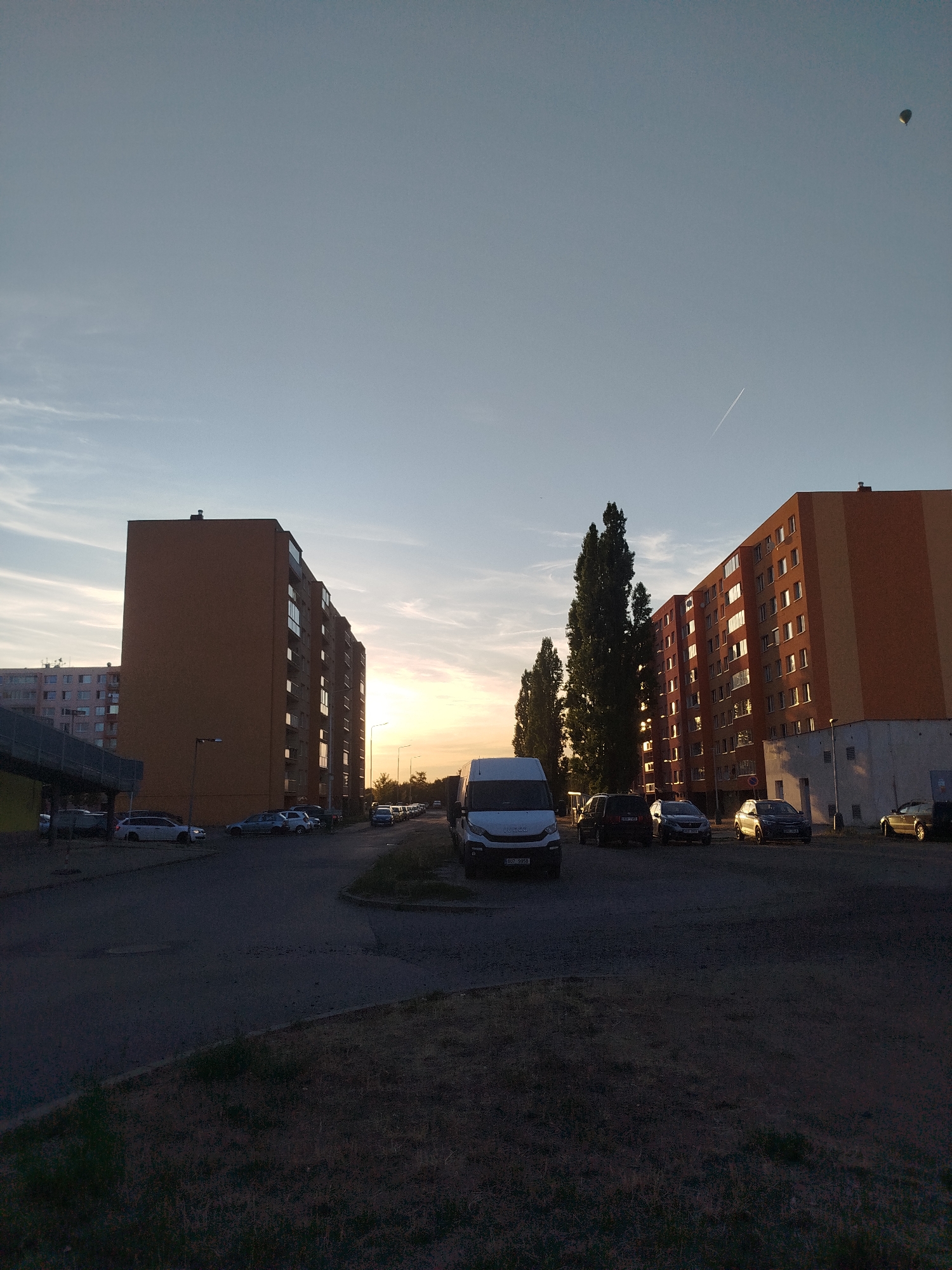 The image shows a street scene at dusk in a city. The sky is a pale blue with some wispy clouds and a contrail from an airplane. A hot air balloon is visible in the upper right portion of the sky. Two large apartment buildings, one with orange siding and the other with a multi-colored facade, are prominent in the background. In the foreground and midground, there are several parked cars, including a white van in the lower left and a dark SUV, a sedan, and a gold car in the midground. Tall, slender trees line the area between the apartment buildings. The ground in the foreground is dark with patches of grass. The overall lighting suggests the sun is setting or has recently set.