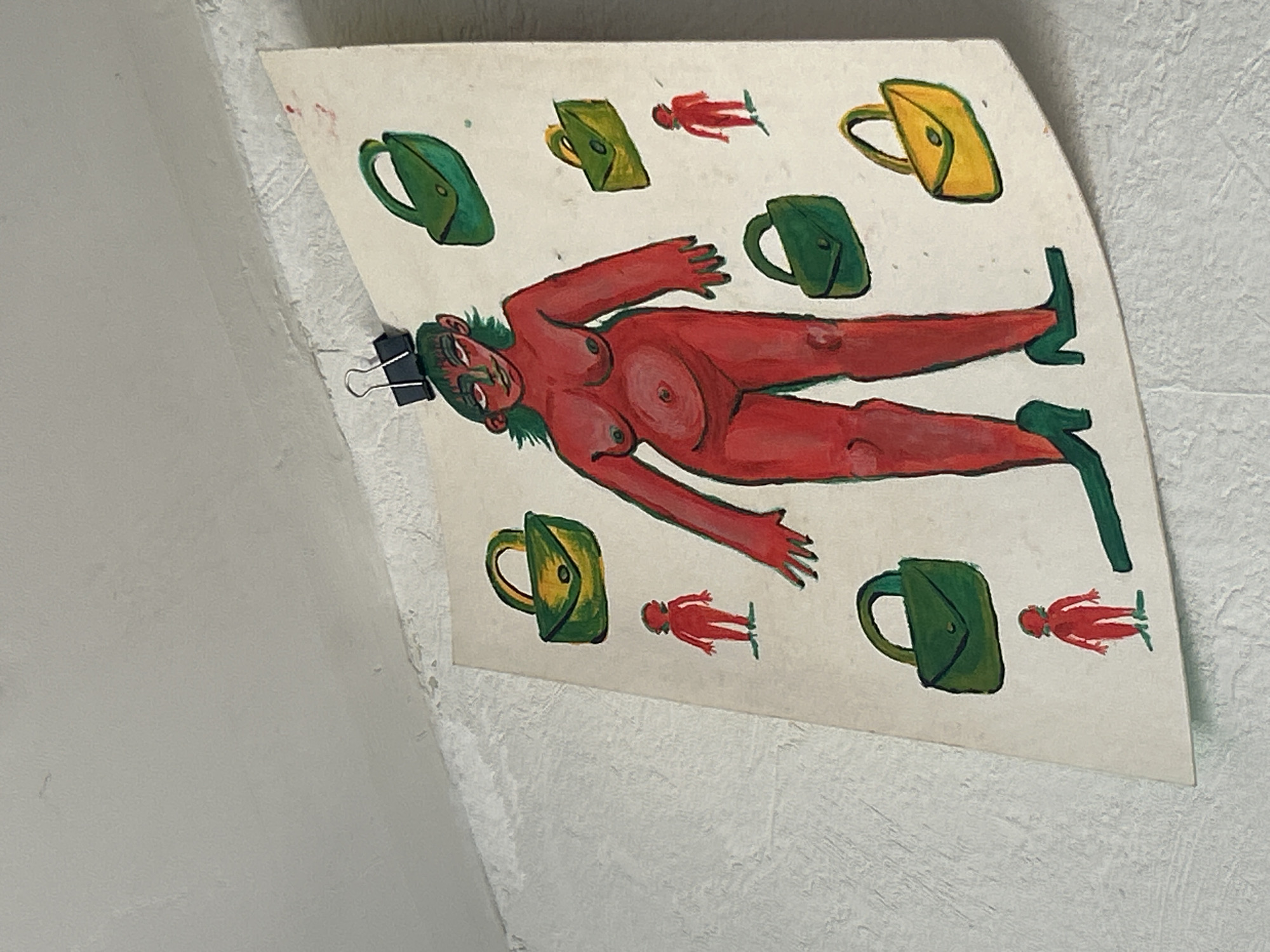 The image depicts a piece of artwork attached to a wall in Unknown, Unknown. The artwork is a drawing or painting on a white, roughly fan-shaped piece of paper. The main subject is a stylized red nude figure with green hair and green pointy shoes, lying horizontally across the paper. The figure is surrounded by several green and yellow depictions of bags or purses and several red human figures. The artwork is attached to the wall by a paperclip or binder clip in the upper left corner. The wall is white with a textured surface.