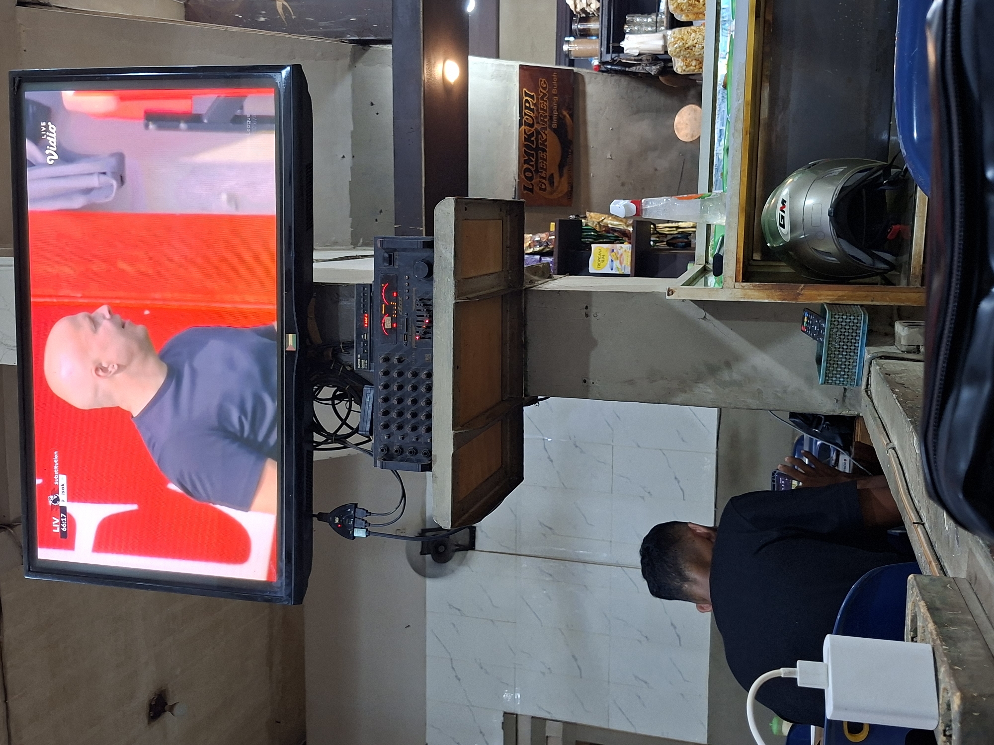 A man is seated, facing right, at a counter in an indoor space, possibly a small business, in the city of Lhokseumawe, Indonesia. He is wearing a black shirt and appears to be looking at something on the counter. Above him, attached to the wall, is a flat-screen television showing a football match, displaying the time 66:17.
To the left of the TV, a sound mixer is mounted on the wall. Various packaged goods, a helmet, and other items are visible on the shelves. A sign that says "LOM KUPI ULEE KARENG Simpang Buloh" is seen above the shelving unit. The walls are tiled, and the overall lighting is moderate. A power adapter and cable are visible near the bottom right of the image.