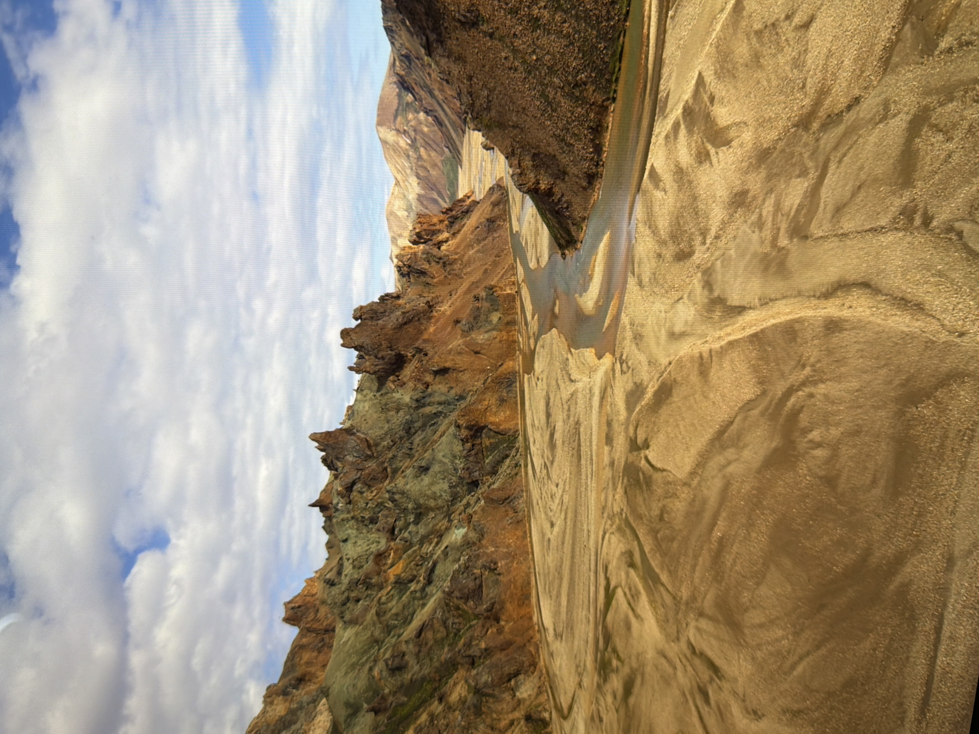 The image displays a dramatic, sloped landscape featuring rugged, rocky hills and a winding, shallow river or stream. The hills on the left are steep and jagged, showing layers of earth and rock in shades of brown, orange, and green, with patches of vegetation. The riverbed, visible on the right, is wide and composed of light-colored sand and gravel, with the water forming a narrow, reflective channel that snakes through it. The textures of the sand appear smooth in some areas and rougher with small pebbles in others, creating patterns shaped by water flow. Above the hills, a vast expanse of sky is filled with fluffy white clouds against a bright blue background. The lighting suggests it is daytime, likely mid-morning or afternoon, with the sun casting shadows that highlight the contours of the terrain. There are no people or man-made objects visible. The scene evokes a sense of natural wilderness and geological formations, with the winding river as a central element of the landscape. The location is identified as Kyiv, Ukraine.