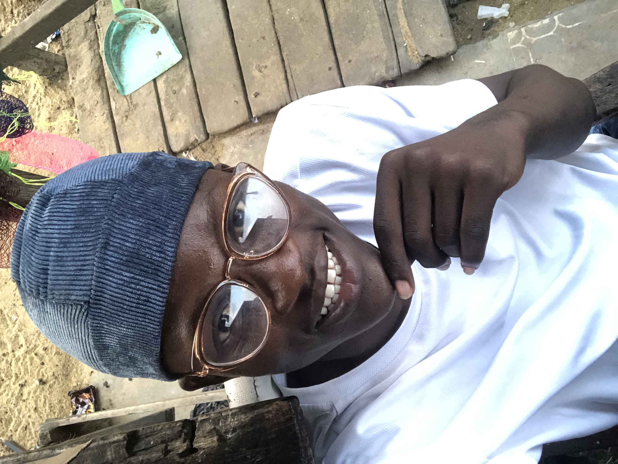 A close-up selfie shows a young Black person wearing a blue ribbed beanie and large, rose-gold rimmed glasses. They are smiling and touching their chin with their right hand, with their fingers resting just below their lower lip. The person is wearing a white t-shirt with a textured pattern. The background suggests an outdoor, somewhat rustic setting, with wooden planks or a walkway and sandy ground. To the right of the person, a light blue dustpan is visible on the wooden planks. The overall lighting is bright, suggesting it was taken during the day. The scene appears to be an informal moment captured in an outdoor area.