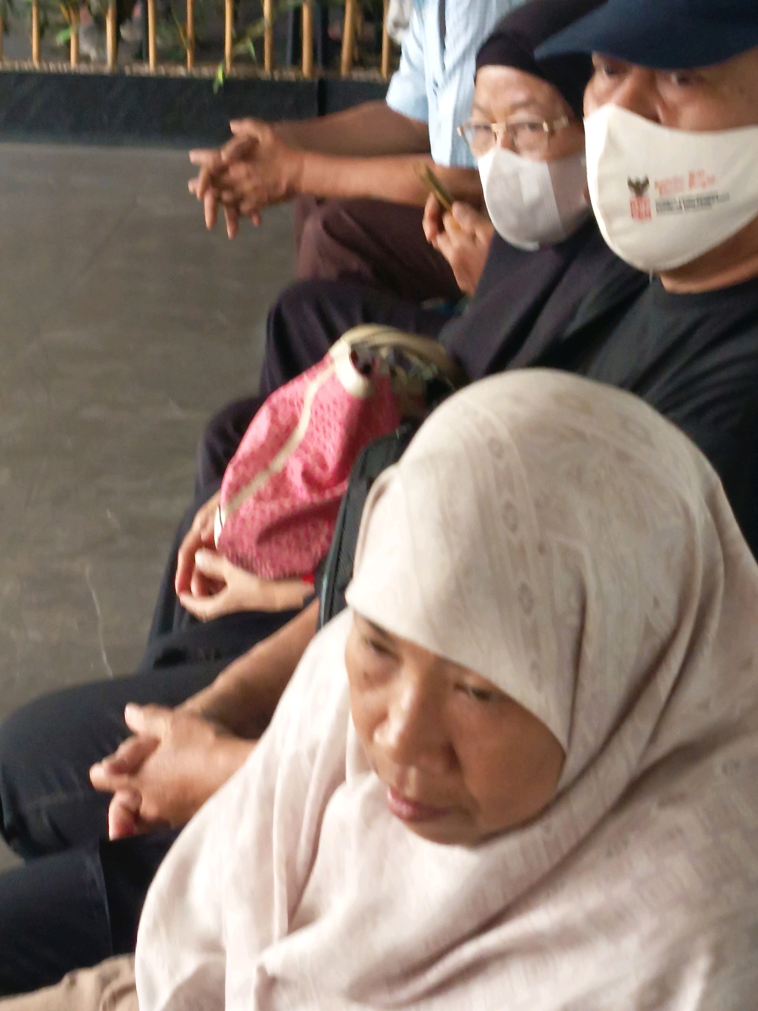 In the city of Tangerang, Indonesia, three individuals are seated, with the most prominent subject being a woman in the foreground wearing a light-colored hijab. Her head is bowed, and her face is partially obscured by the fabric. To her left, another person, also wearing a hijab and glasses, is holding a smartphone. Further left, a third individual, seen from the shoulders up, is wearing a blue collared shirt. The background consists of a dark, textured surface and a blurred wooden fence with vertical slats. The overall scene appears to be an indoor or covered outdoor waiting area.