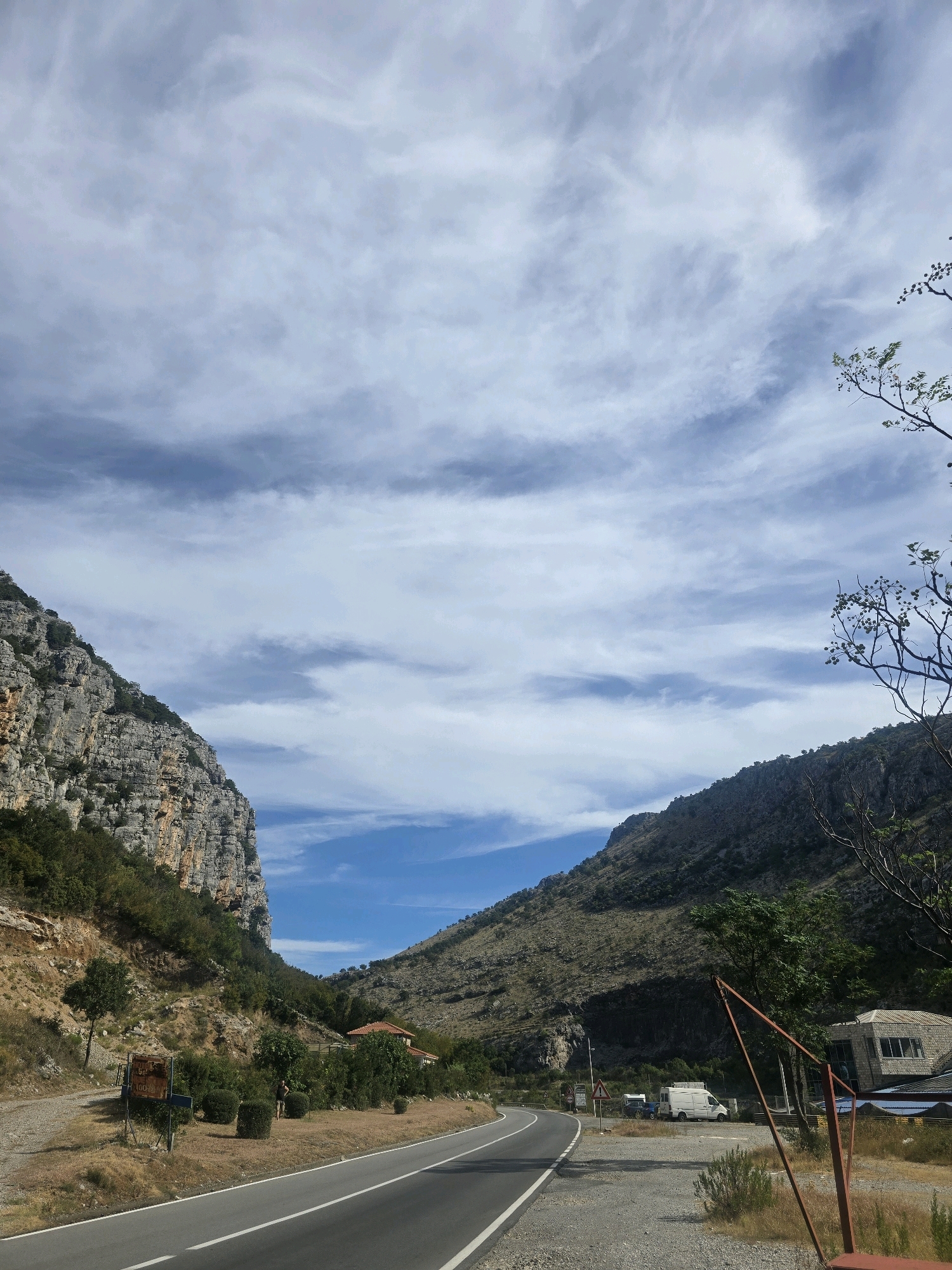 A paved road curves through a mountainous landscape under a partly cloudy sky. The road is bordered by dry, sparse vegetation and gravel on the right and a steep, rocky cliff face dotted with trees on the left. In the middle distance, a few trees and a red-tiled roof house are visible amongst the greenery. Further along the road, a white van and a blue car are parked near a stone building with a glass facade. A person wearing dark shorts and a t-shirt stands near the house. A rusted metal structure, possibly part of a gate or fence, stands in the foreground on the right. The scene is likely located in a rural or semi-rural area near Podgorica, Montenegro.