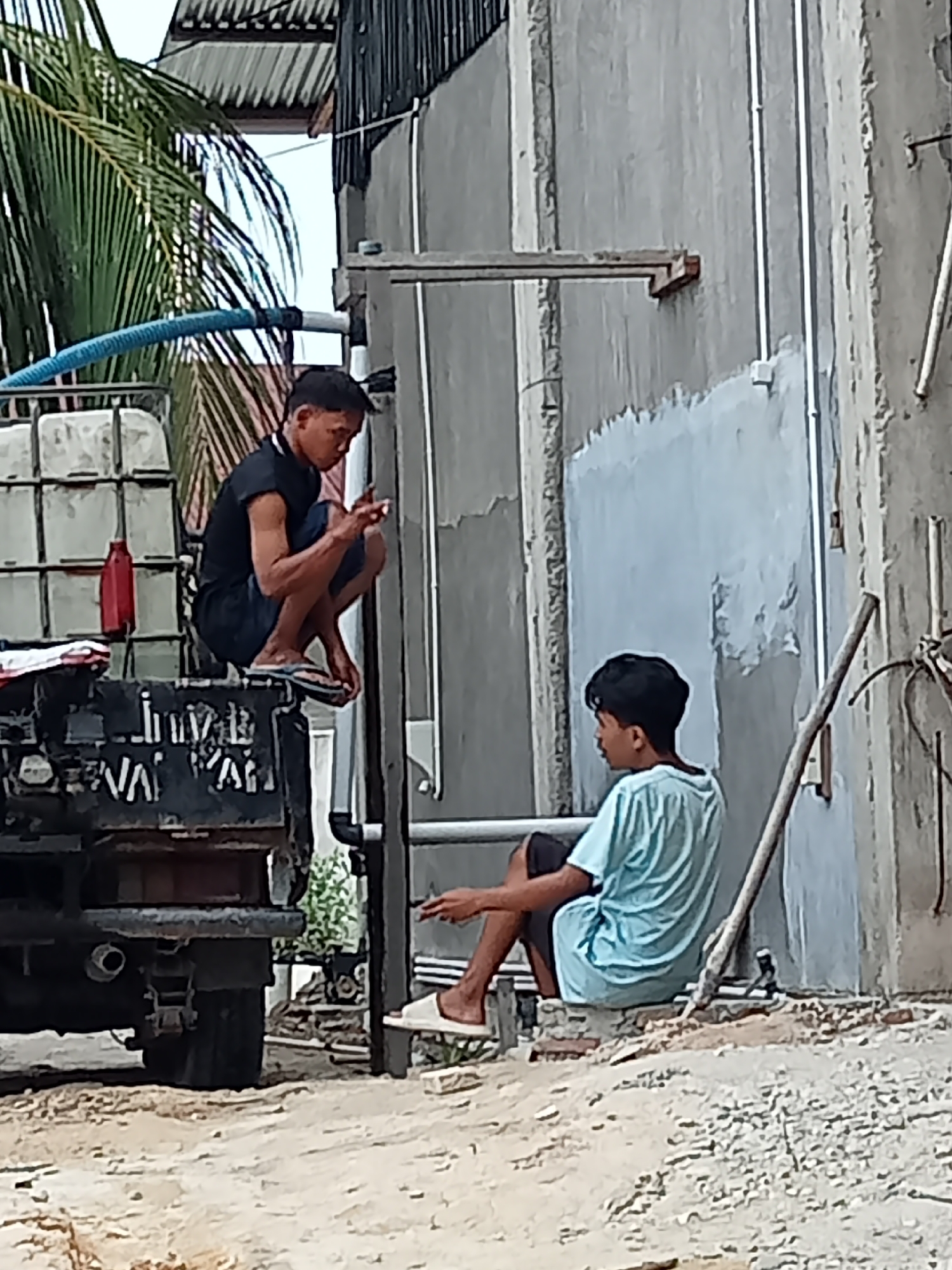 Two young males are seated outdoors, on what appears to be a sunny day, in a location that could be Melayu Besar Kota, Indonesia. One male, wearing a black t-shirt and dark shorts, is crouched on the back of a dark-colored truck. He is holding a small object, possibly a cigarette or a phone, in his right hand. The other male, wearing a light blue t-shirt and dark shorts, is seated on a low, light-colored ledge or structure. He is also holding a small object in his right hand. The truck has a large white container or tank on its bed, and a blue hose is connected to it. Behind the individuals, there is a rough, light-gray wall, possibly part of a building under construction or renovation, with visible piping and scaffolding. A palm frond is visible in the upper left of the frame. The ground is a mixture of sand and dirt, with some loose gravel. The overall scene suggests a casual or resting moment during work or an outdoor activity.