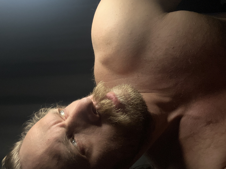 An extreme close-up shot focuses on a man's face from a low angle, set against a dimly lit background. The man has fair skin, light-colored hair, and a short beard. His eyes are visible, appearing to be slightly open. The lighting emphasizes the contours of his face and neck. Behind the man's head, a portion of his upper chest and shoulder is visible, suggesting a bare torso. The overall composition creates a sense of intimacy and vulnerability.