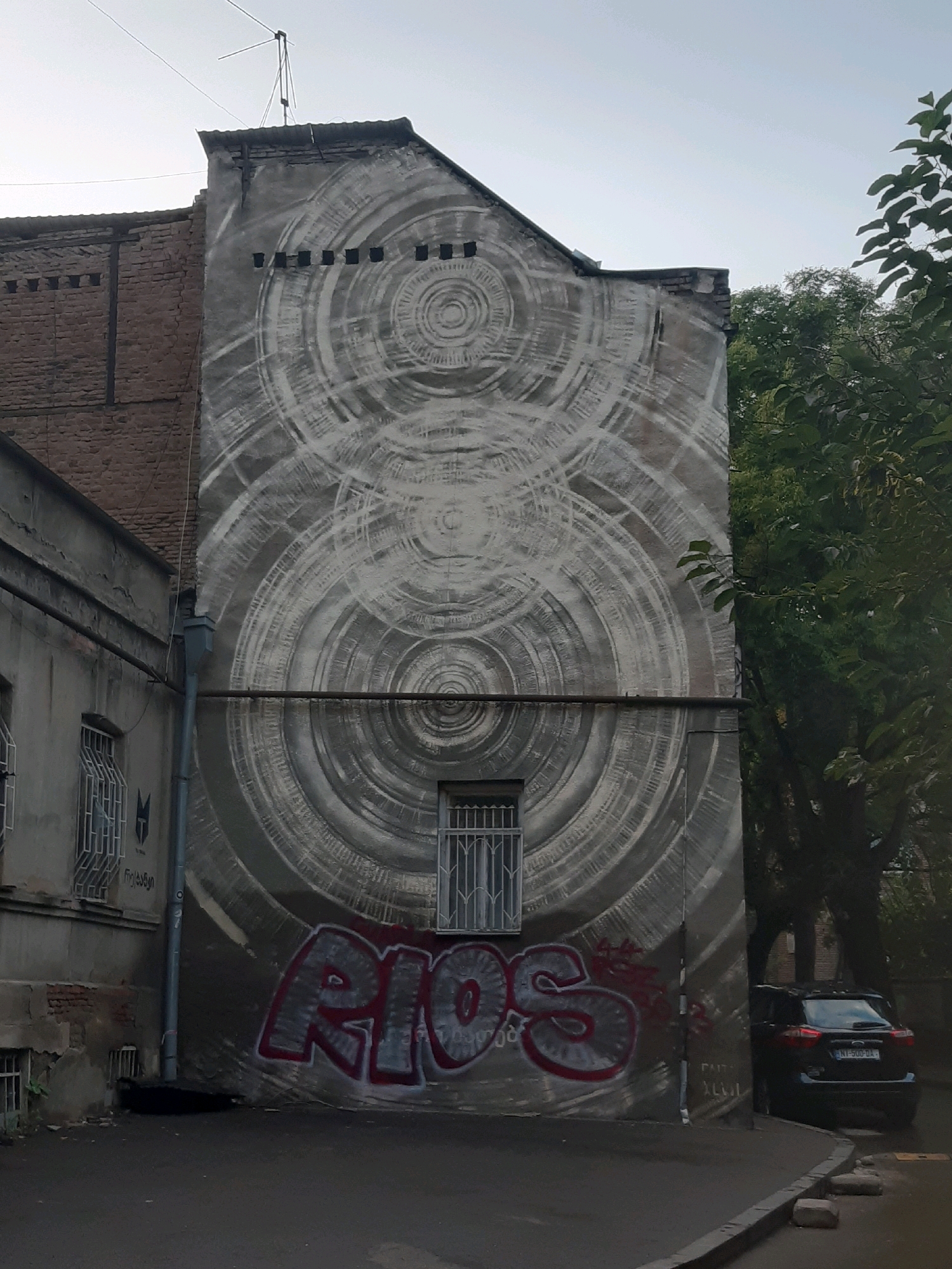 The image shows a mural on the side of a building in Tbilisi, Georgia. The mural features a large, abstract design of concentric circles and lines in shades of gray. The building itself is made of brick and concrete, with several windows and a drainpipe. There is graffiti spray-painted in red and gray on the lower part of the wall, spelling out "RIOS". A black car is parked on the street in front of the building, and trees and foliage are visible on the right side of the image. The sky is overcast.