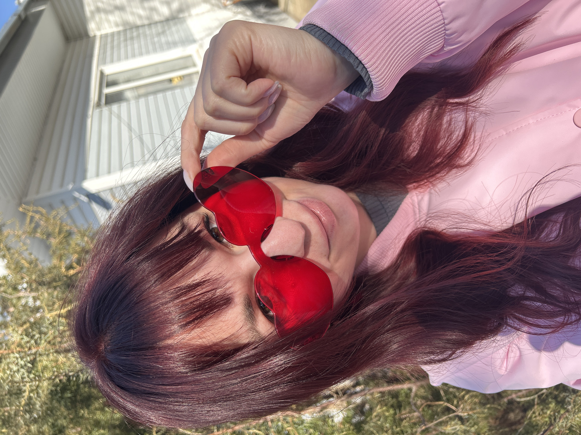 A woman with red hair and bangs is holding red heart-shaped sunglasses in front of her face. She is wearing a light pink jacket with a gray undershirt. The background consists of a white building with a window and green trees or bushes. The setting is outdoors, likely in a city environment given the presence of a building. The specific location of the image in the city of Фоки, Russia cannot be determined from the visual content alone.