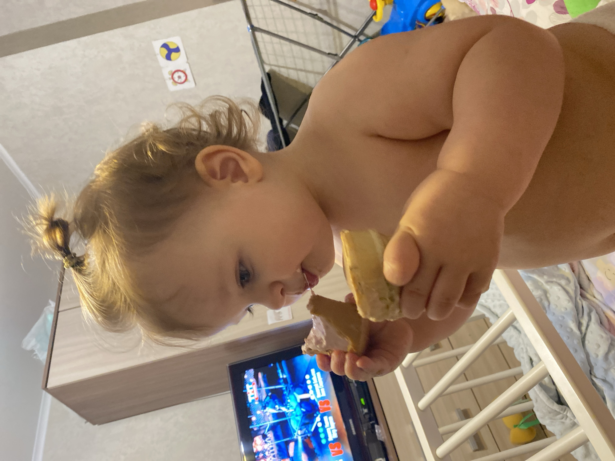 A toddler stands indoors, eating a sandwich, in front of a television showing a wrestling match. The location is Unknown, Unknown. The toddler has light skin and blonde hair pulled into a small ponytail. The TV sits on a cabinet, and behind the toddler is a white crib. On the wall, two stickers are visible. Clothes hang on a drying rack in the background. The toddler is holding the sandwich with both hands, apparently taking a bite.
