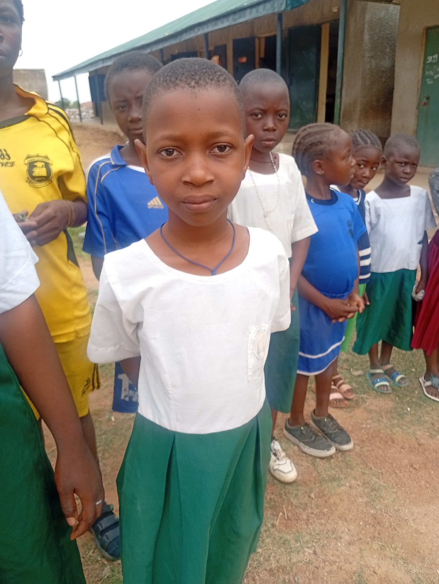 A group of children stand outdoors in what appears to be Narayi, Nigeria. The children are wearing a variety of clothing, including school uniforms in blue, yellow, white and green. The ground is bare and dry. A simple building with a green roof is in the background.