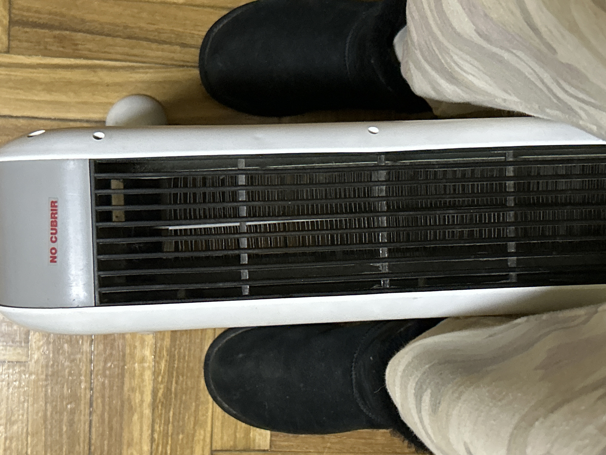 An overhead, wide shot shows a person's legs in black suede boots and patterned loungewear positioned around a white tower heater. The heater is positioned vertically and has a black grill with silver elements visible behind it. The text "NO CUBRIR" in red is visible at the top of the heater. The heater is placed on a light brown, wooden floor with visible plank lines. The scene appears to be indoors.