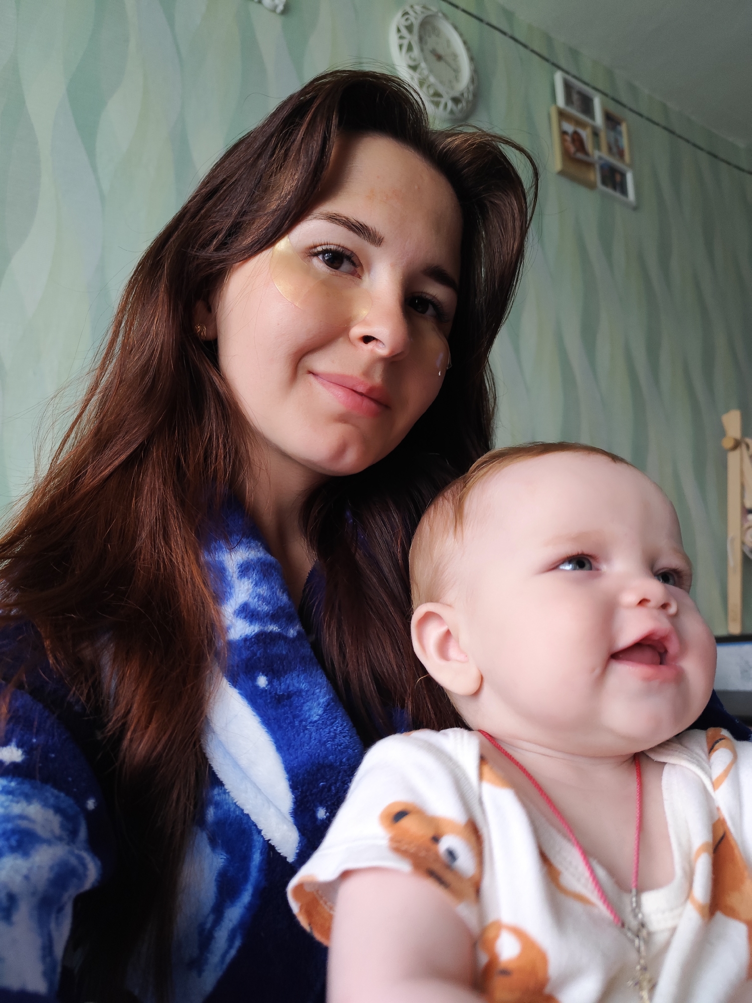 The image depicts a woman holding a baby. The woman has long brown hair and appears to be wearing under-eye patches. She is wearing a blue and white robe. The baby is fair-skinned with reddish hair and is wearing a white onesie with a bear pattern. A red string necklace with a cross pendant is visible around the baby's neck. The background includes a green wall with a subtle wave pattern, a clock, and several framed pictures. Based on the surroundings, the location seems to be inside a home. No evidence in the image indicates that this photo was taken in the city of Фоки, Russia.