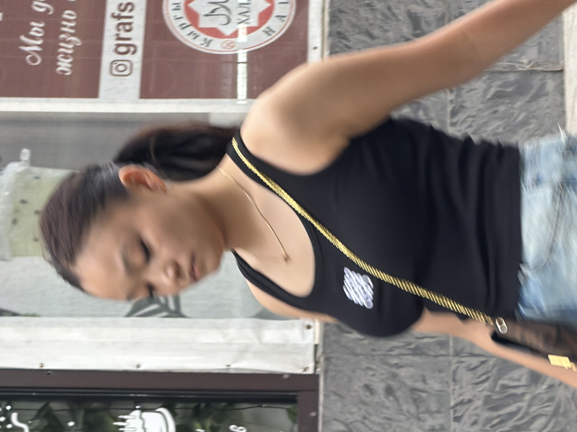 A young Asian woman with dark hair in a ponytail walks down a street, her head turned to her left. She is wearing a black tank top with a white logo on the front and a gold chain necklace. A brown and gold patterned purse with a gold strap is slung across her chest. She is also wearing light-wash denim shorts. The background shows a wall with signage, including a circular emblem with text and a logo that appears to be an Instagram handle. The ground in front of her is paved with gray stone.