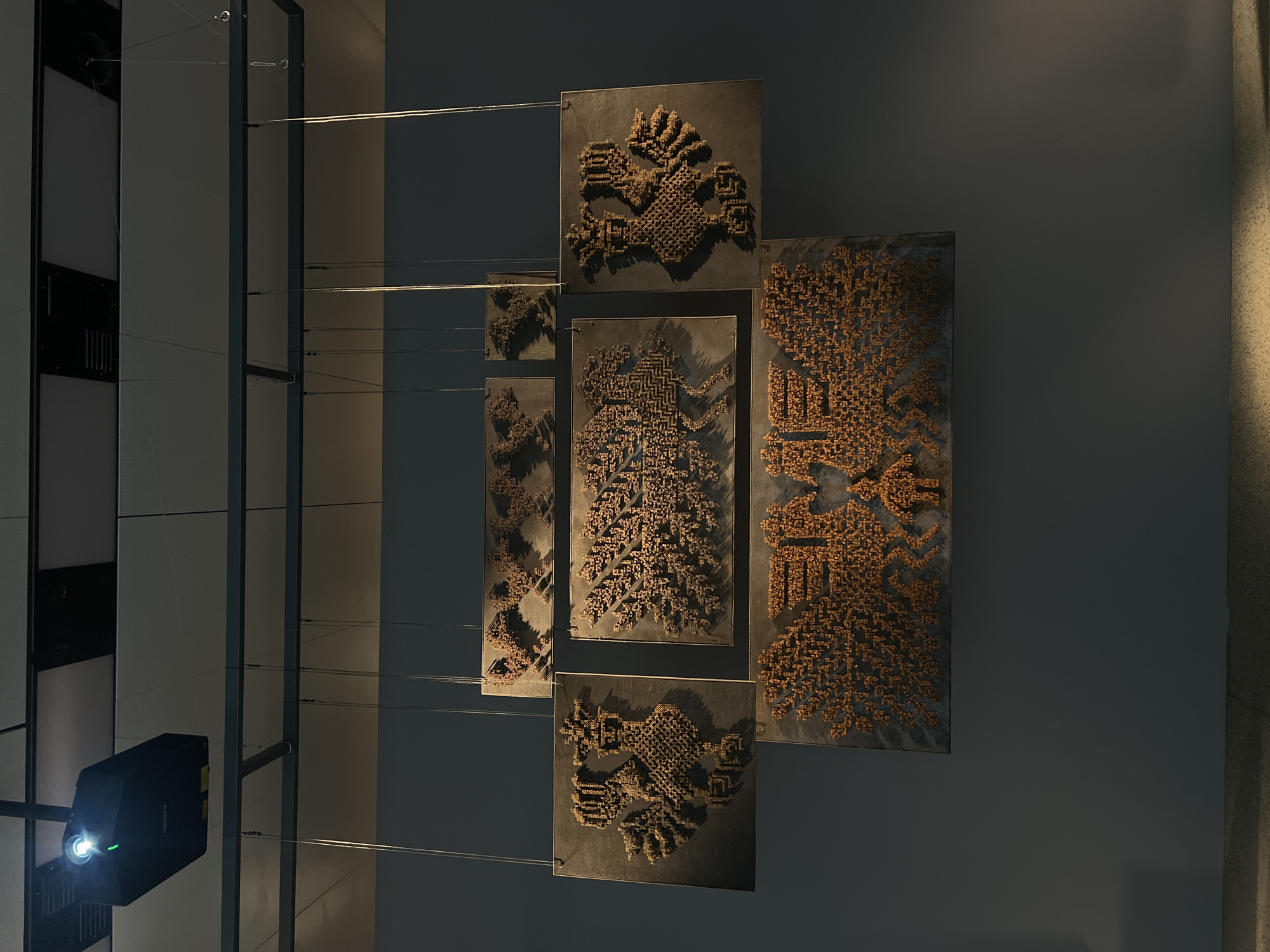 The image captures an art installation hanging from the ceiling of a museum or gallery space. The installation consists of five square or rectangular panels arranged in a cross-like formation. Each panel displays an intricate design made of what appears to be small, uniform, brown or bronze-colored components. The designs resemble stylized figures or patterns, reminiscent of ancient glyphs or geometric motifs. Thin cables or wires suspend the panels from a structural grid above. To the left of the installation, a projector is mounted on a vertical support structure, with its light emitting. The walls are plain and neutral in color, with a gradient effect suggesting a light source from the right. The precise location and subjects of the art are indeterminate from the image.