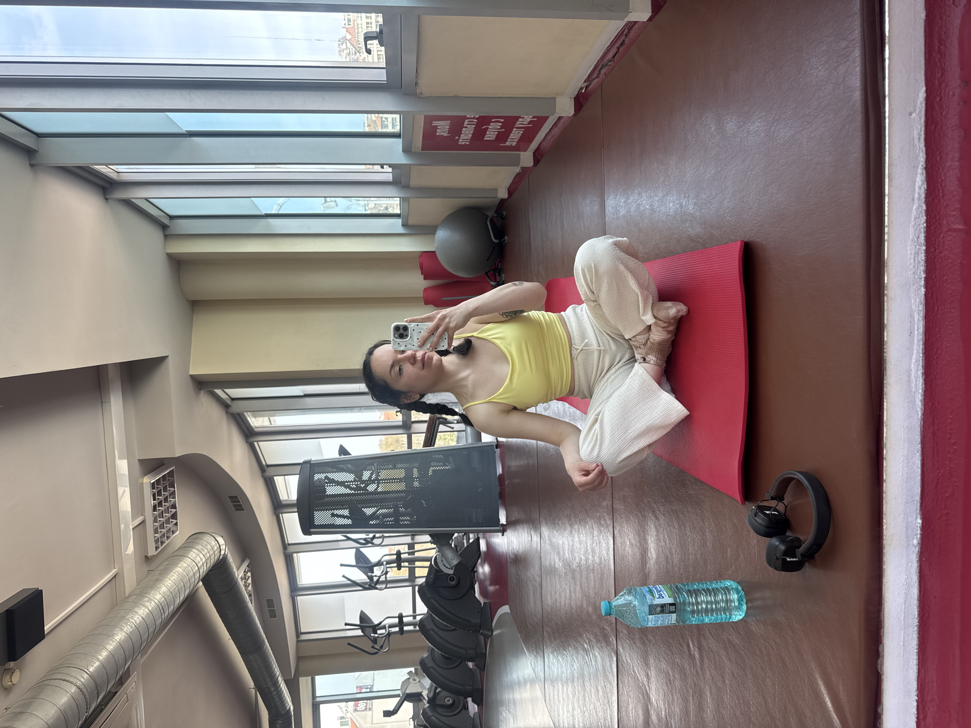 A woman sits in a cross-legged position on a red yoga mat, holding a cell phone in her right hand as if taking a picture. She is wearing a yellow tank top and white pants. A water bottle and black headphones are on the floor near the yoga mat. Gym equipment is visible in the background. A large window shows the city of Burgas, Bulgaria.