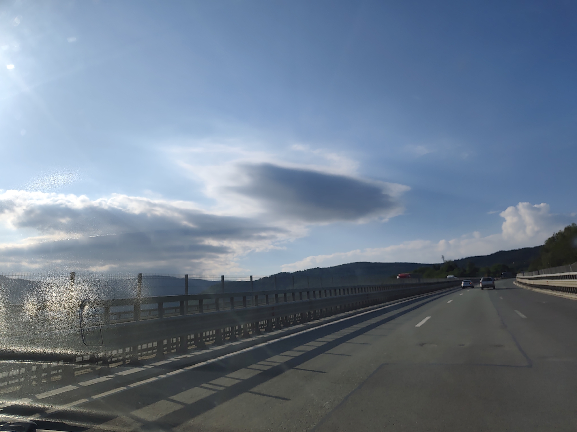 The image captures a view from a vehicle traveling on a highway in Vrachesh, Bulgaria. The highway has multiple lanes and is bordered by a metal guardrail and a fence. The sky is a bright blue with scattered clouds, and the sun is visible in the upper left corner, casting lens flares and creating a bright sheen on the vehicle's windshield. In the distance, rolling hills covered in vegetation can be seen. Several cars are on the highway, and shadows from the guardrail stretch across the asphalt. The overall scene suggests a scenic drive on a sunny day.