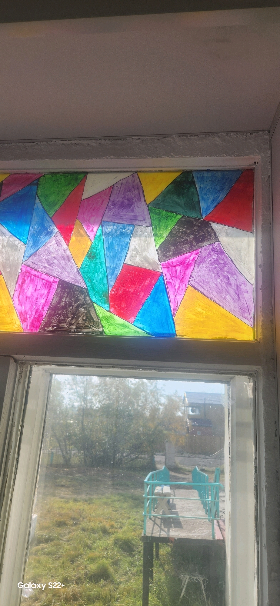 The image depicts a stained-glass-like window decoration featuring a geometric pattern of colorful triangles, made with what appears to be colored paper or transparency sheets. The decoration is mounted in a window frame, with the lower portion of the image showing a view through the window of an outdoor scene. The outdoor scene includes overgrown grass, trees, a concrete platform, and a turquoise-colored railing with a bench and table on it. In the background, a building with a distinct turret-like structure can be seen, suggesting it might be a church or a similar establishment. The timestamp on the lower left corner indicates "Galaxy S22+", suggesting the photo was taken with a Samsung Galaxy S22+ smartphone. While the image does not explicitly show the city of Nyurba, Russia, the presence of a building with a Russian architectural style in the background and the mention of the location in the prompt suggest this is the context. The overall scene suggests a domestic setting, likely a home with a view of a yard or a public space.