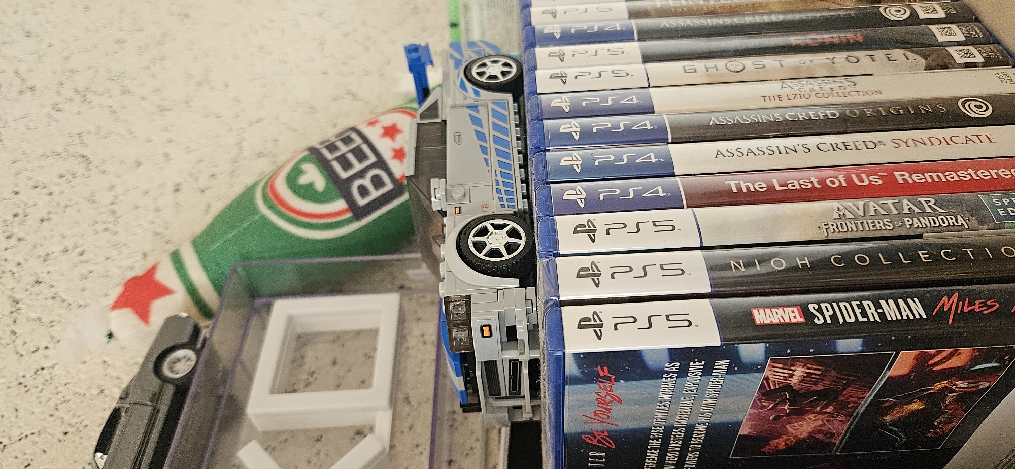 The image shows a stack of PlayStation 4 and PlayStation 5 game cases, including titles like "Assassin's Creed Origins," "The Last of Us Remastered," and "Marvel's Spider-Man: Miles Morales." The game cases are arranged vertically and are partially obscured by a toy car and a green beer bottle cover with red stars. The background is a light-colored surface. The location is the city of Sofia in Bulgaria.