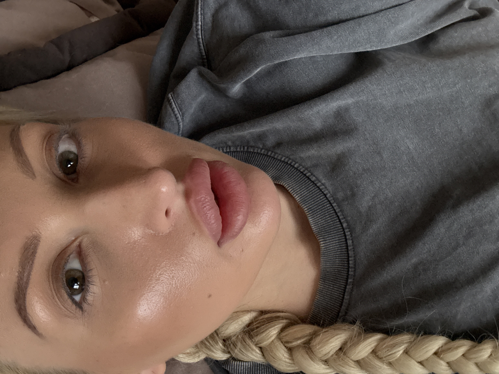 A close-up image depicts a person with fair skin, brown eyes, and blonde braided hair. They are wearing a gray sweatshirt with a ribbed neckline. The person appears to be lying down on a gray surface. The location and city are unknown.