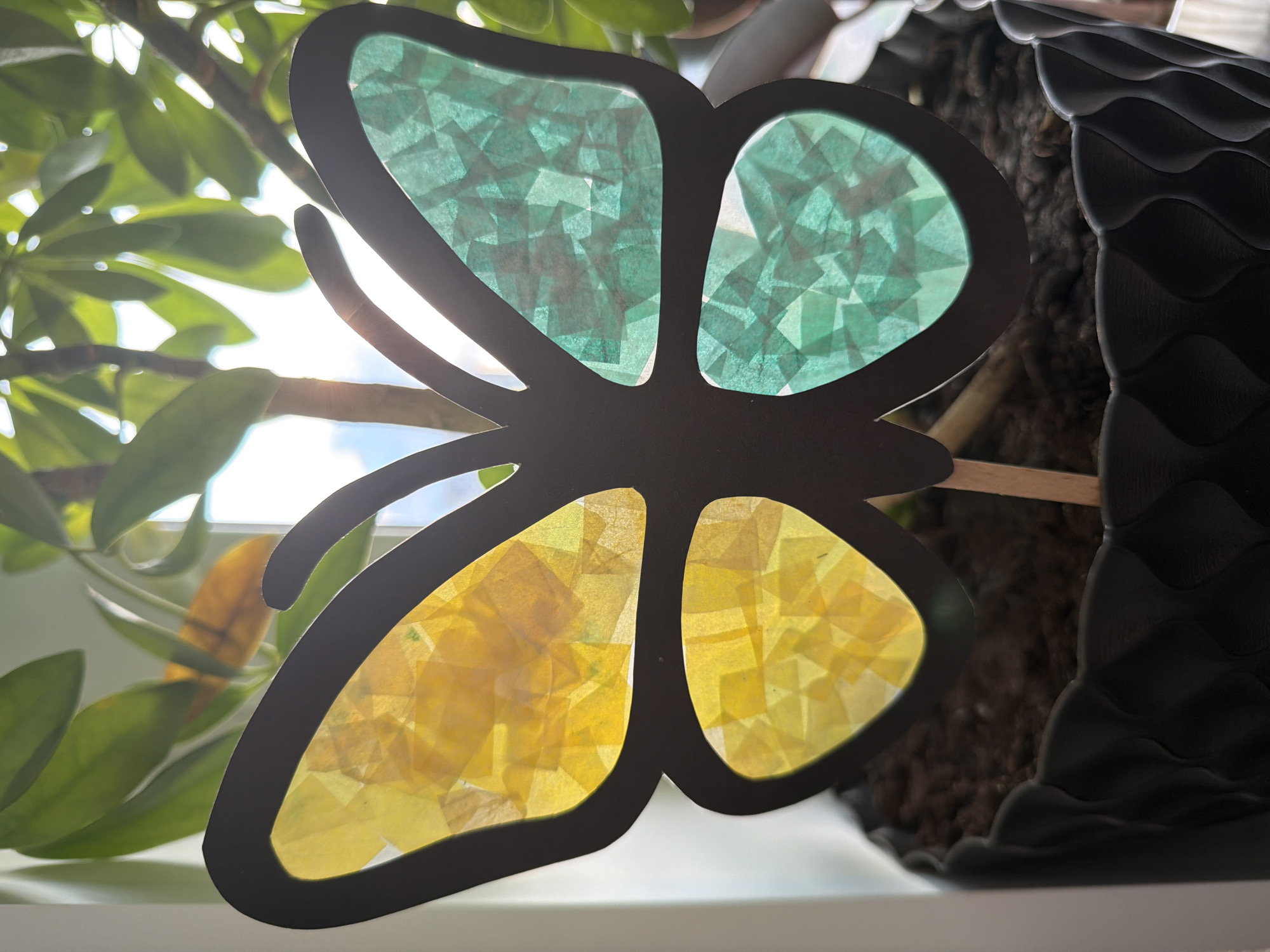 A close-up, eye-level shot shows a black silhouette of a butterfly decoration with yellow and green tissue paper on its wings. The butterfly is stuck in the soil of a black, textured planter. Behind the butterfly, a large green plant with sunlight filtering through its leaves is visible. The background is blurred, with hints of a bright sky and window frame. The scene appears to be indoors, possibly near a window, in a residential or office setting. There is no indication of the specific location within the city of Yekaterinburg, Russia.