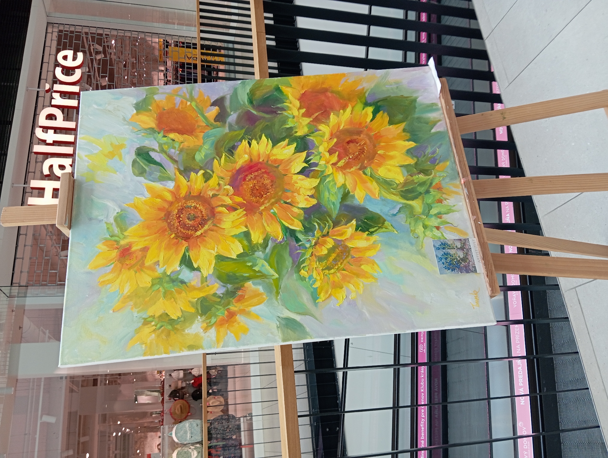 An oil painting of sunflowers is displayed on a wooden easel in front of a storefront with a "HalfPrice" sign. The painting is on a canvas and is placed at an angle, facing slightly to the right. The sunflowers are rendered in bright yellow and orange tones with green foliage against a light blue and white background. A small sticker is affixed to the lower right corner of the canvas, and the artist's signature, "Torresko," is visible in the lower left corner.
The background reveals a shopping mall environment. In the distance, through a glass partition, a retail space is visible with displays of clothing and accessories, including backpacks and hats. The storefront's metal security gate is partially lowered. The lower part of the image shows a black metal railing and a white tiled floor. Pink signage with text in Slovak is visible on a lower level through the railing, suggesting it is part of a commercial area. Specifically, signs read "NOVÁ PREDAJŇA NA PRÍZEŇ" (New store for favor) and "VOŇAVÝ DOMOV" (Fragrant home), indicating commercial establishments.
The presence of the "HalfPrice" signage and the interior retail displays strongly suggest the location is within the OC Promenada shopping mall in Nitra, Slovakia. The overall scene captures an art display within a commercial setting. There are no people visible in the immediate vicinity of the painting.