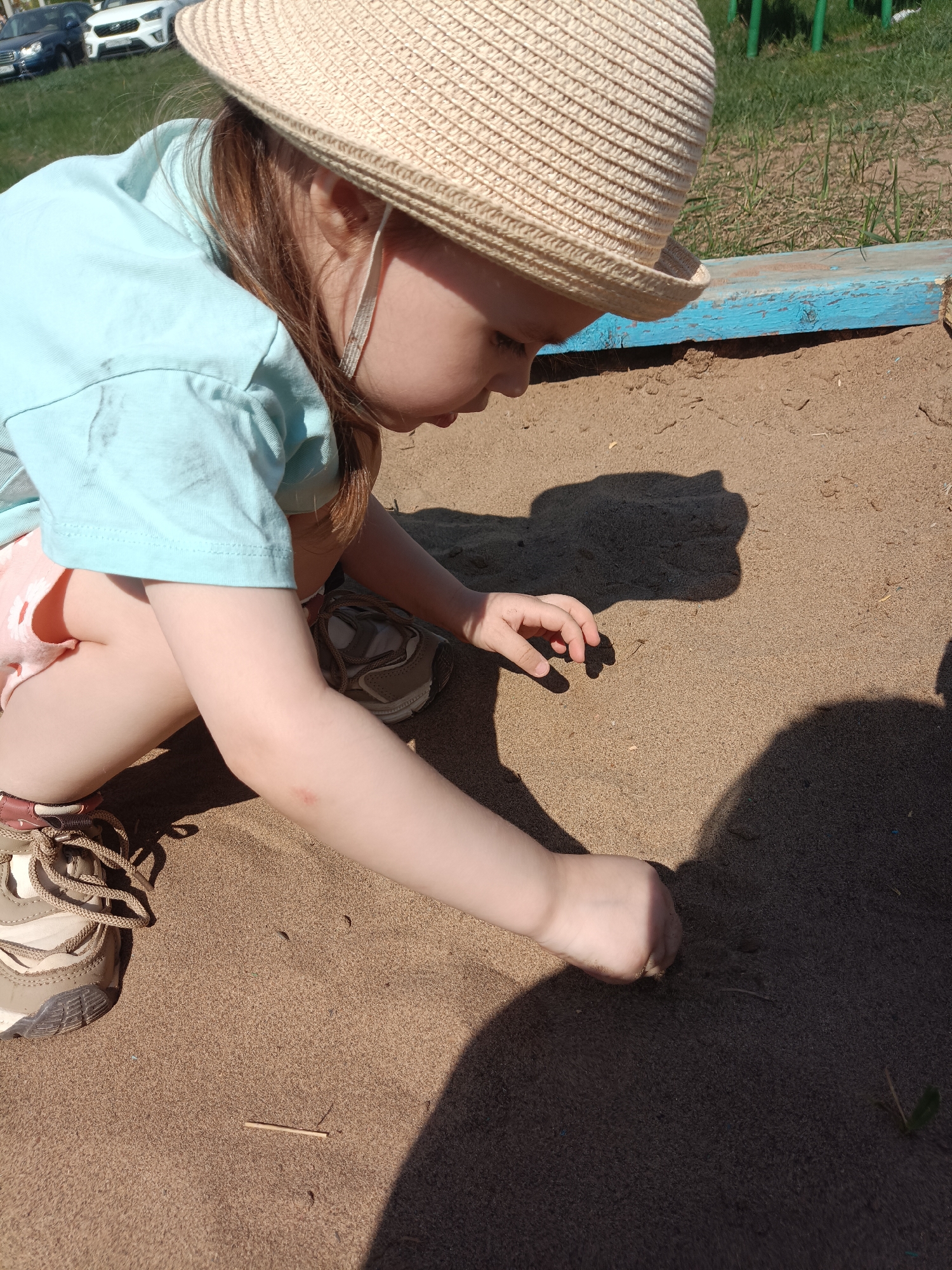 A young child wearing a straw hat, a light blue shirt, and pink shorts is sitting in a sandpit. The child is using their hand to touch or dig in the sand. The location is identified as Noviy, Russia by geolocation data. In the background, there is a blue painted border to the sandpit, some green grass, and parked cars. The scene is bathed in sunlight, casting shadows from the child and their surroundings onto the sand.