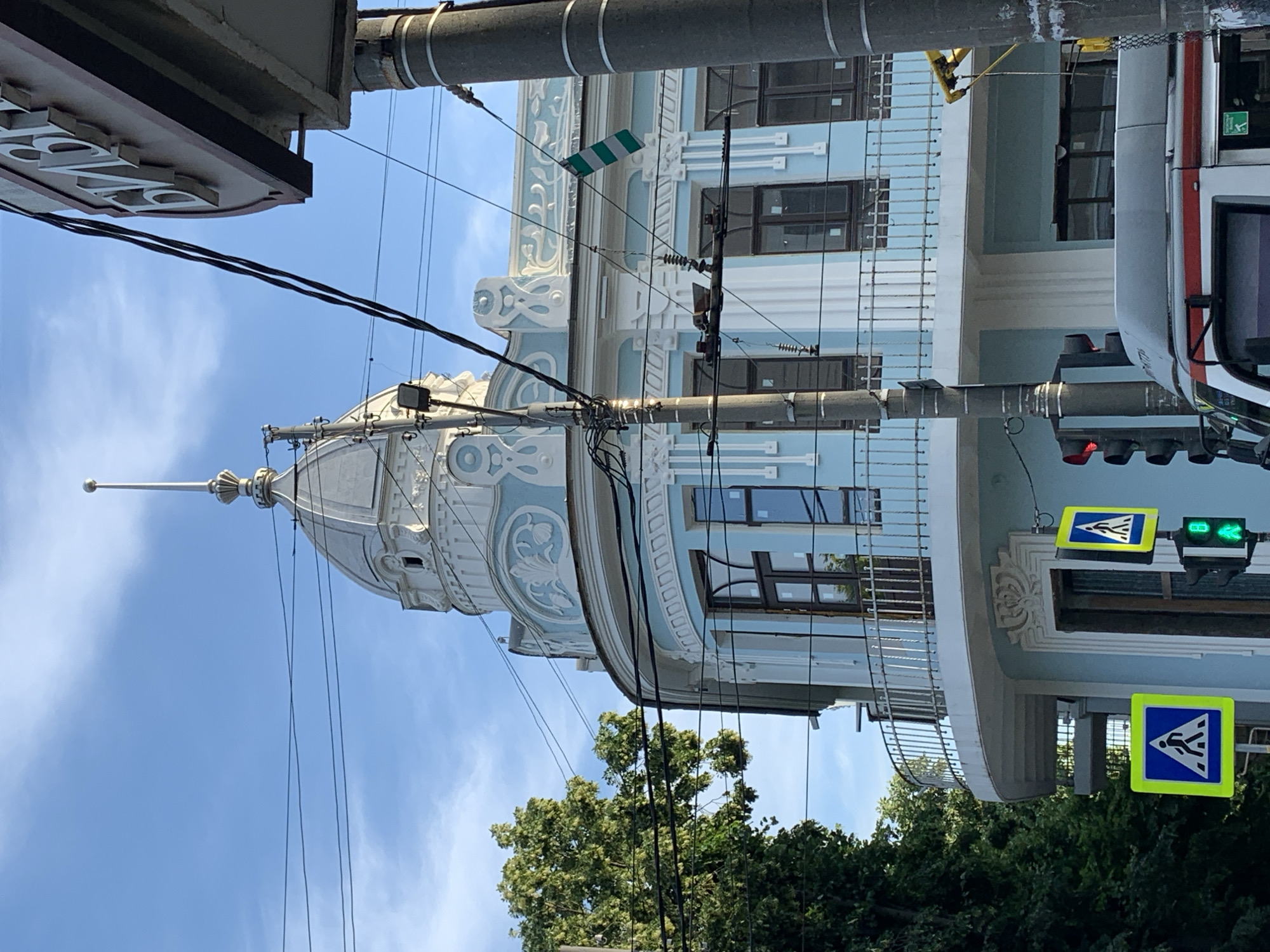 The image shows a light blue building with ornate white trim and a domed roof topped with a spire. A curved balcony with a metal railing is visible on the building. In the foreground, there are traffic lights and pedestrian crossing signs. Overhead, a network of electrical wires crisscrosses the sky. To the left, lush green trees partially obscure the building. A portion of a sign with white lettering on a dark background is visible on the right side of the frame. A vehicle, possibly a tram, is partially visible at the bottom of the image. The sky is clear and blue.