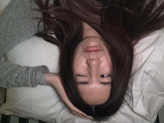 A female individual is depicted from a high angle, lying supine on white bedding in an indoor setting. Her long, dark brown hair, parted down the middle, is spread across a white pillow. She presents a winking expression; her right eye is closed, and her left eye is partially open, looking towards the right side of the frame. Her lips are slightly parted, and a small silver ring is visible in her right nostril. An arm, covered by a grey, textured long-sleeved garment, extends from the right side of the frame with its hand resting gently on the subject's head. Crinkled white sheets or duvet covers are visible in the left foreground, while a dark vertical element, consistent with a bed frame or headboard, is present in the upper right background. The image contains no visual elements indicating a specific geographical location, such as Le Havre, France.