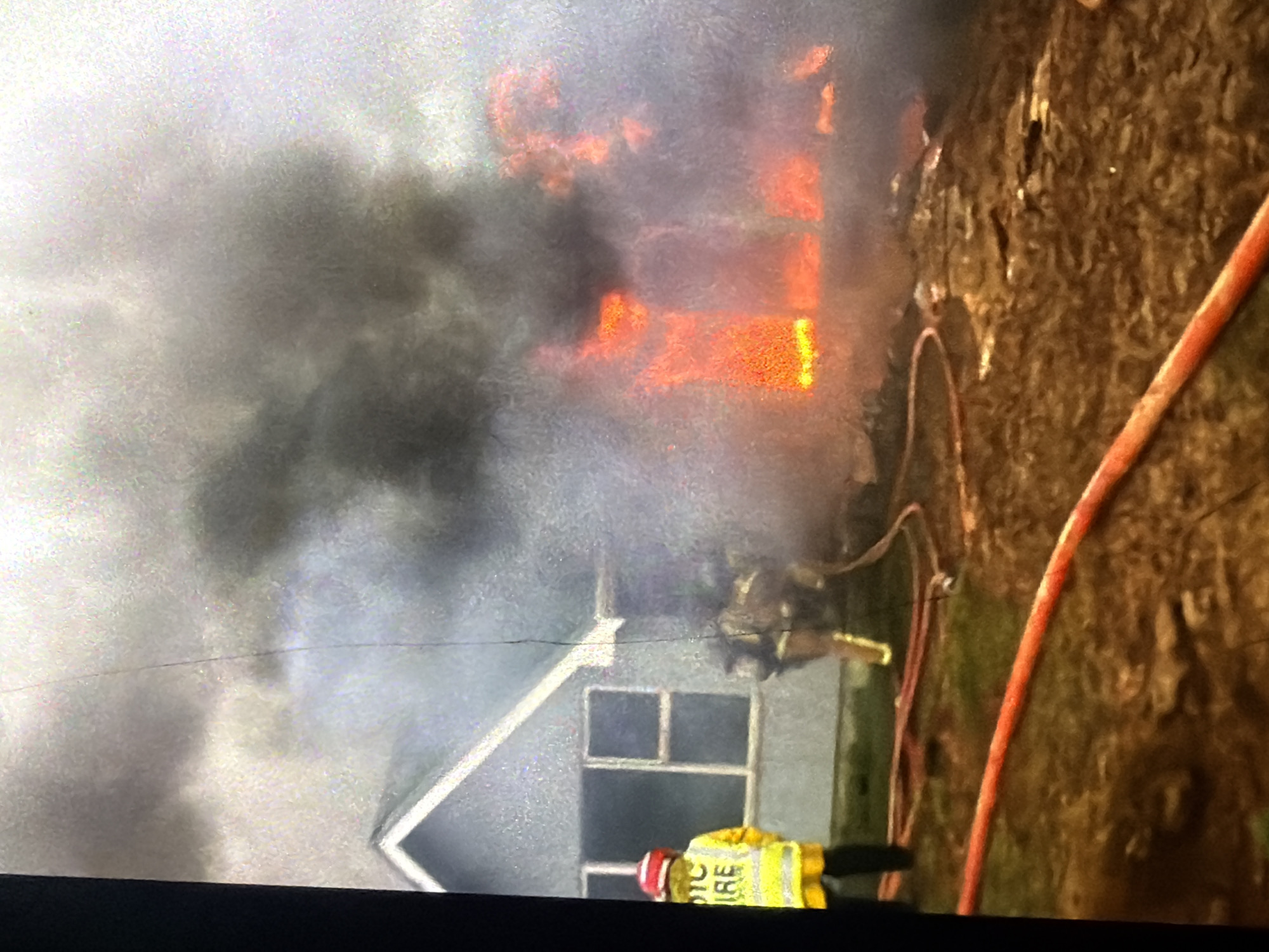 In the city of Bukuru, Nigeria, a house is engulfed in flames. Thick, dark smoke billows from the structure, obscuring much of the scene. Two firefighters in full gear are actively engaged in combating the fire. One firefighter, positioned on the left side of the frame, is wearing a bright yellow jacket with "FIRE" visible on the back and a red helmet. This firefighter is facing away from the camera, towards the burning house. Another firefighter, also in protective gear, is closer to the house, partially obscured by smoke and flames. Red fire hoses are laid out on the ground, connecting to a water source and extending towards the blaze, indicating an ongoing firefighting operation. The environment appears to be a residential area with the house showing visible signs of fire damage. The ground is covered in debris and dark soil.