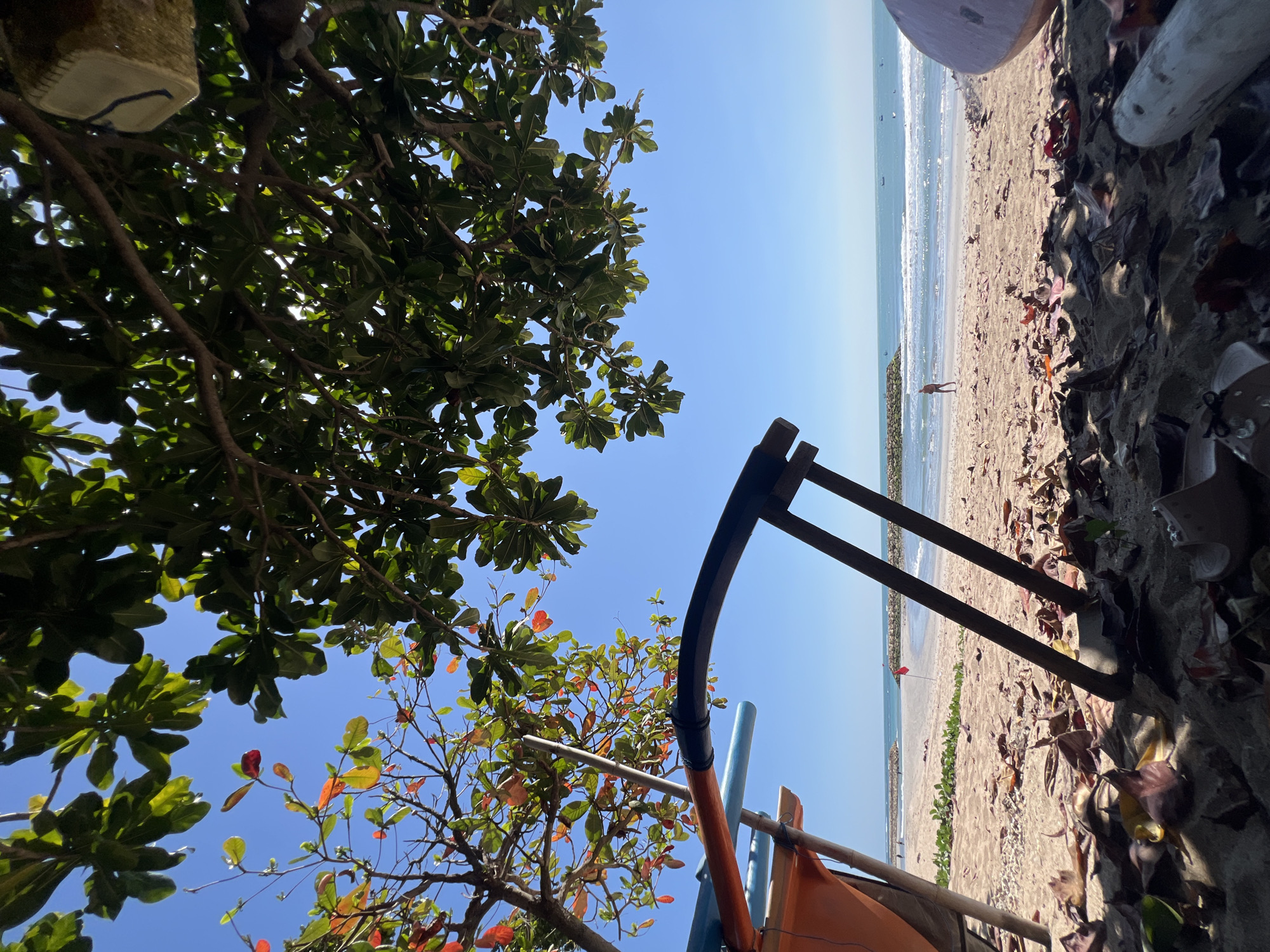 The image depicts a low-angle view of a beach scene in Kuta, Indonesia, under a clear blue sky. In the foreground, fallen leaves are scattered on the sand, partially obscuring a light-colored object and the base of what appears to be part of a boat or shelter structure with wooden supports and orange and blue elements. Further back, the sandy beach stretches towards the ocean, with gentle waves breaking on the shore. Two rocky breakwaters extend into the water, and a red flag is visible on one of them. A single person is walking on the beach in the mid-ground. The ocean extends to the horizon, with several small boats visible on the water. Lush green foliage from trees overhangs the scene from the top, with some leaves showing hints of red and orange. The overall impression is a serene beach environment.