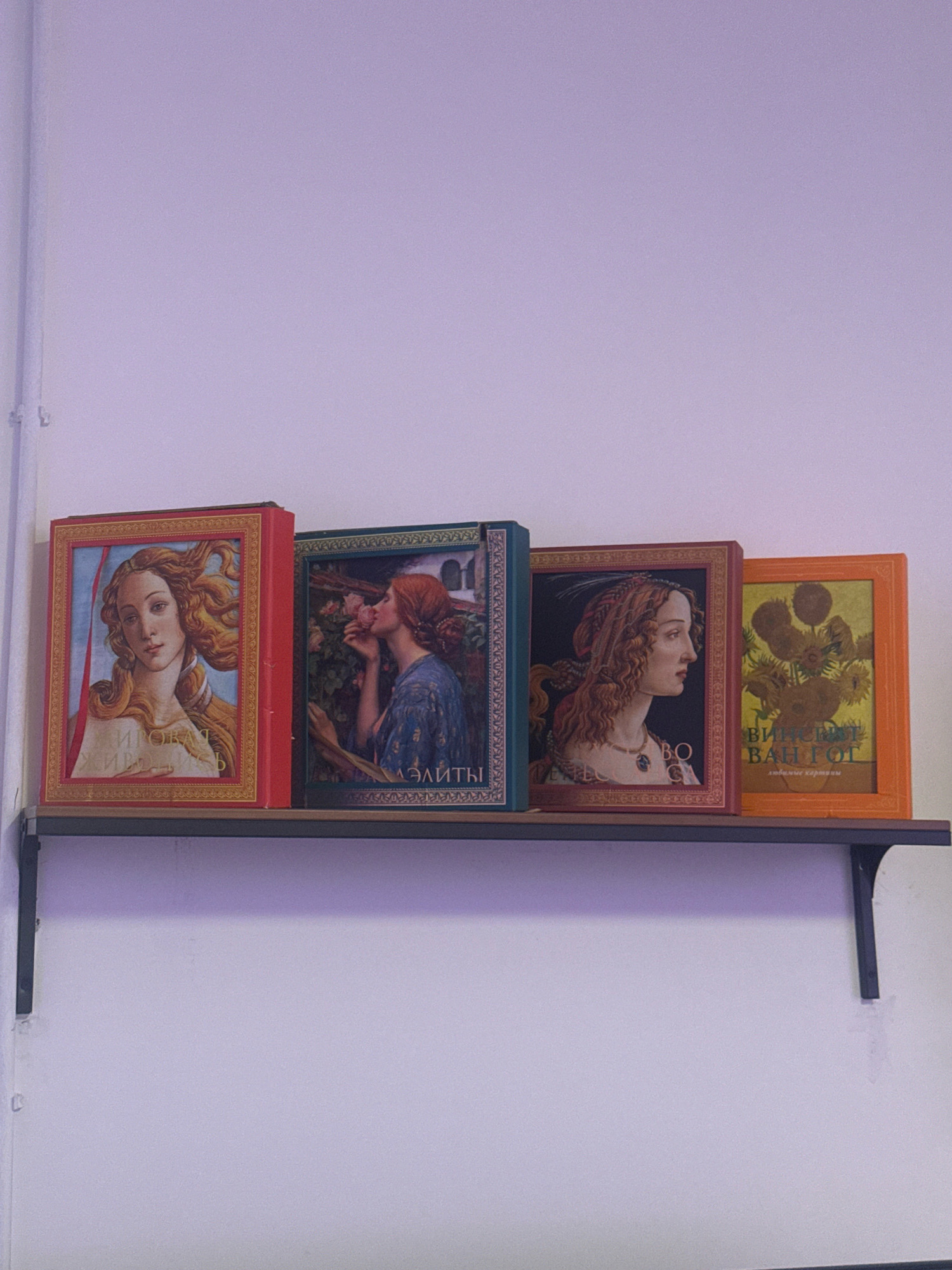 A shelf with four art books is mounted on a light purple wall. The books are arranged in a row from left to right. The first book on the left is red and features a painting of a woman with flowing hair. The second book is dark green and displays a painting of a woman smelling a rose. The third book is maroon and shows a profile portrait of a woman with curly hair. The fourth book, on the far right, is orange and features a painting of sunflowers. The shelf is made of dark wood and is supported by black brackets. The overall scene appears to be indoors. The city of Germasogeia Municipality, Cyprus, is not identifiable from the provided image.