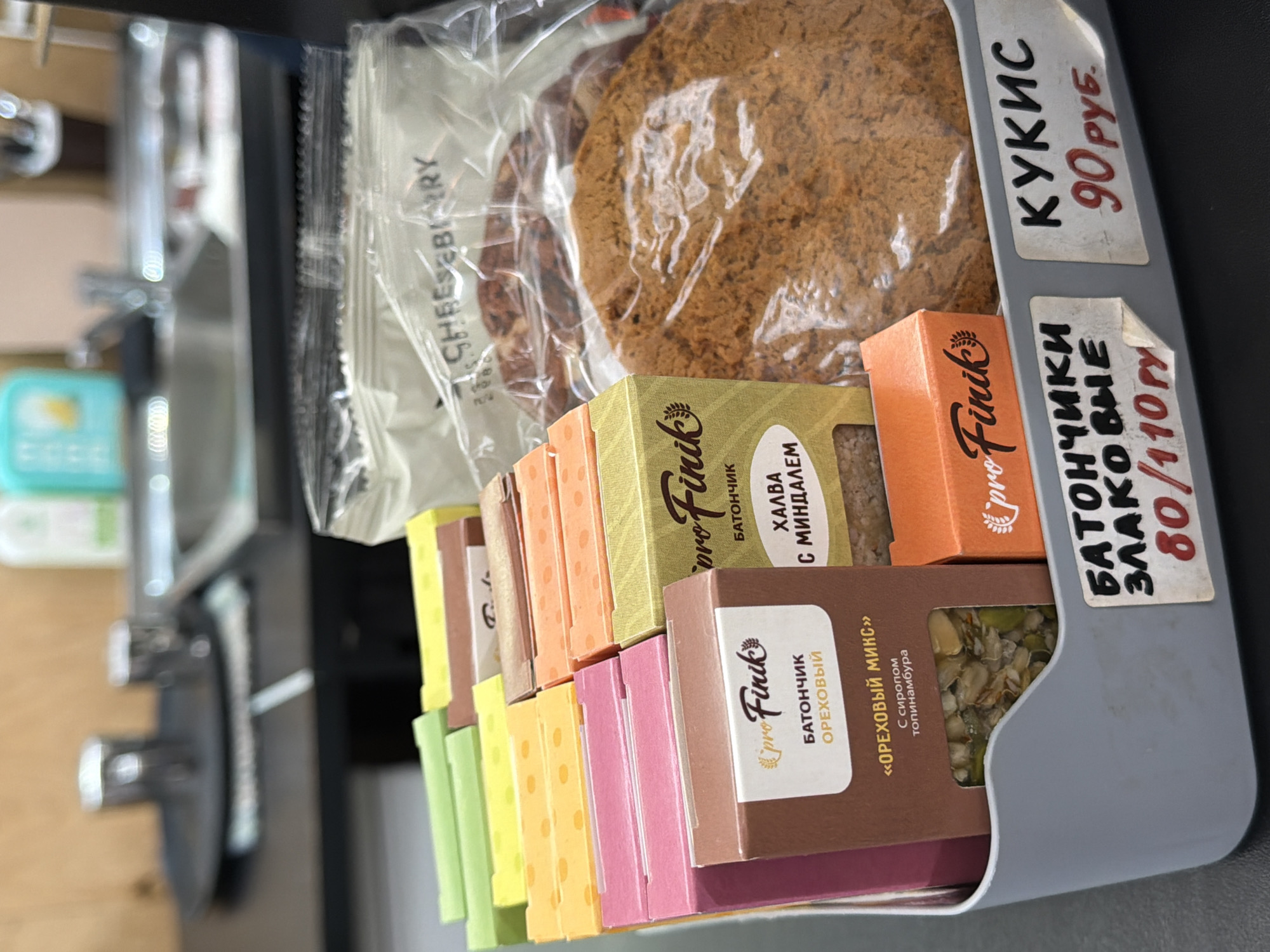 The image shows a close-up of a selection of packaged snacks and treats displayed in a gray container, likely for sale. The snacks include individually wrapped cookies labeled "CHEESEBIARY" and individually boxed snack bars labeled "pro Finik." The "pro Finik" bars appear to be available in various flavors, including "OREKHOVY" (nutty mix) and "KHALVA s MINDALEM" (halva with almonds). Price tags indicate that "KUKIS" (cookies) cost 90 rubles, while "BATONCHIKI ZLAKOVYE" (cereal bars) cost 80/110 rubles. The overall setting seems to be a store or café in Chaikovsky, Russia, given that the labels are in Russian.