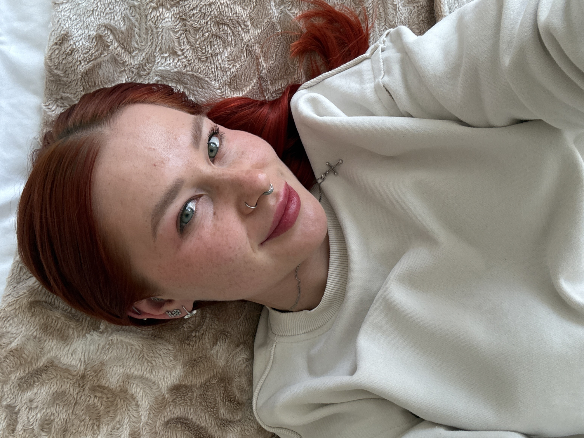 A fair-skinned woman with auburn hair and freckles lies on a bed covered with a textured tan blanket. She is wearing a light-colored sweatshirt. Her head is tilted slightly towards the camera. She has facial piercings including a nose ring and multiple earrings. A delicate silver chain necklace is visible. Location cannot be verified.