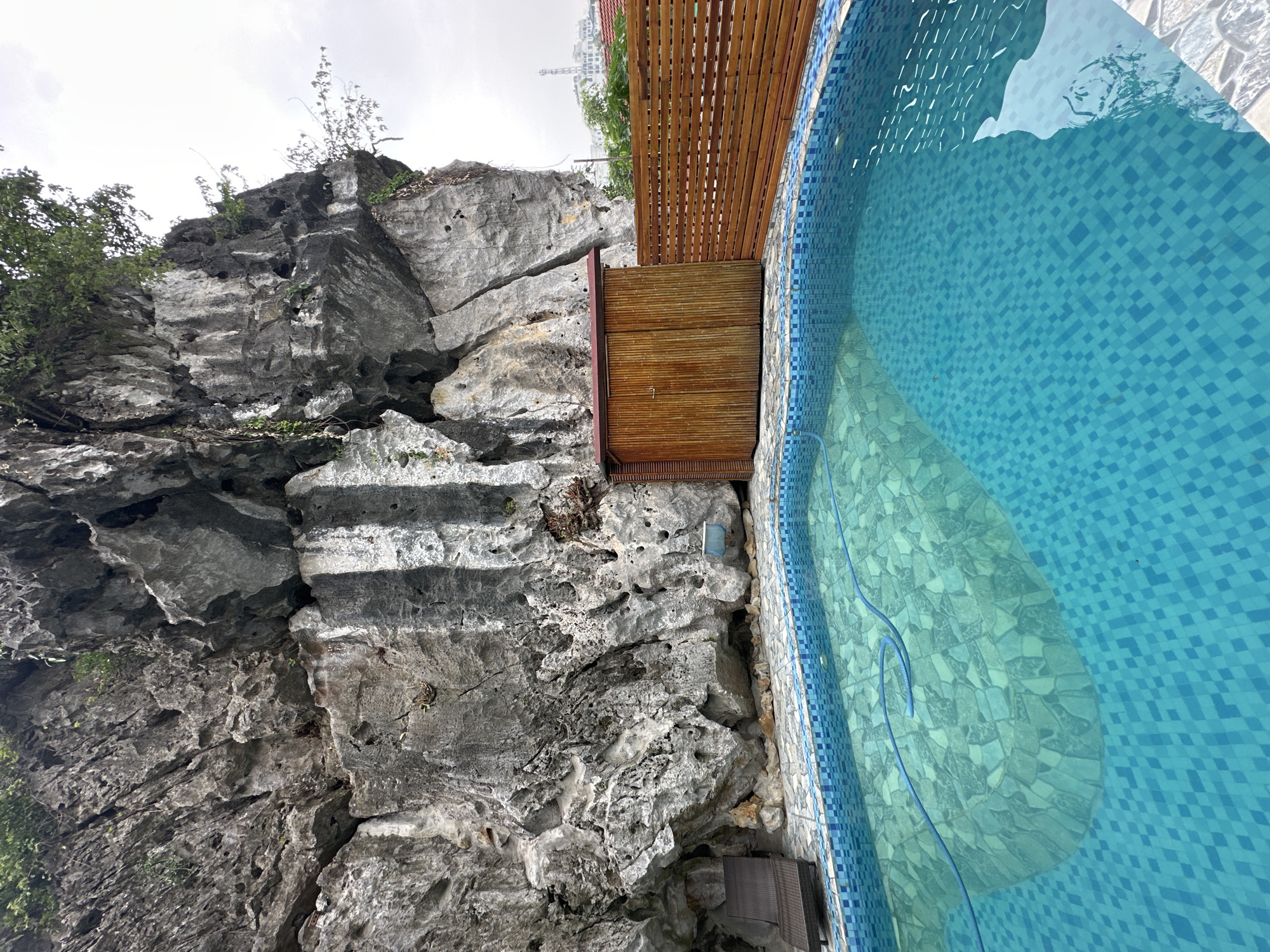 The image depicts an outdoor swimming pool situated in front of a large, rugged rock face. The pool is filled with clear turquoise water and is tiled with small blue and white squares. A blue hose lies coiled at the bottom of the pool. To the right of the rock face, there is a small, wooden shed with a reddish-brown roof and a wooden fence. A lounge chair is visible to the left of the rock face, near the pool's edge. In the background, beyond the fence and shed, buildings and trees can be seen under an overcast sky. The scene appears to be in a resort or private residence. The general area is likely Nam Hoa Lư Ward, Vietnam, known for its karst landscapes. There are no people visible in the image.