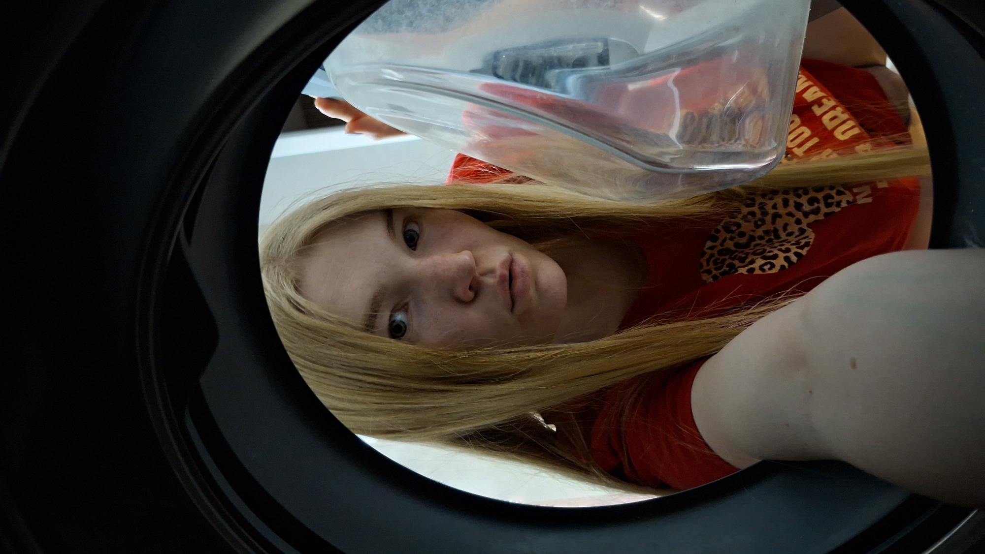 A young, fair-skinned woman with long blonde hair is depicted in a close-up, low-angle shot from inside what appears to be a black, circular container, possibly a bin or large pipe. She is looking directly into the "camera," her face slightly tilted. She wears a red t-shirt with leopard print design and the text "TOO DREAM" visible. A clear, plastic container is positioned above her, partially obstructing the view. The location is unknown; there is no clear indication that the image was taken in Chaikovsky, Russia.