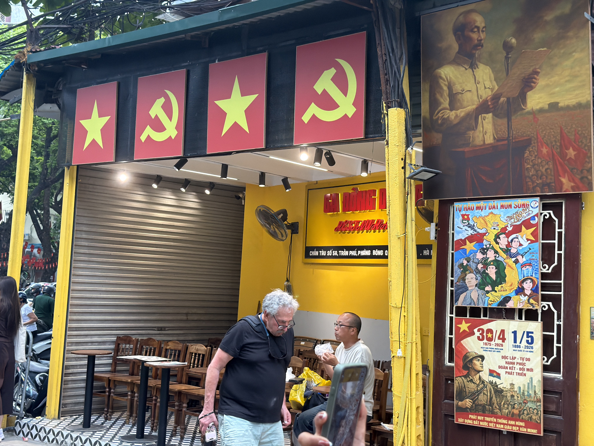 The image shows a street scene in Hà Nội, Vietnam, featuring a building with political iconography. The building has four red panels above the entrance, alternating between a yellow star and a yellow hammer and sickle. Above this, there is a painting of Ho Chi Minh giving a speech. To the right of the entrance, there are two posters, one featuring illustrations and maps, and the other with a soldier and civilians. The signage on the yellow building displays the address "CHẮN TÀU SỐ 5A, TRẦN PHÚ, PHÒNG BÔNG Q". A middle-aged man in a black t-shirt and light-colored shorts stands holding a bottle, next to another man with glasses who is sitting and eating food. A hand holds up a smartphone as if taking a picture of the scene. There are tables and chairs nearby and motorbikes parked on the street.