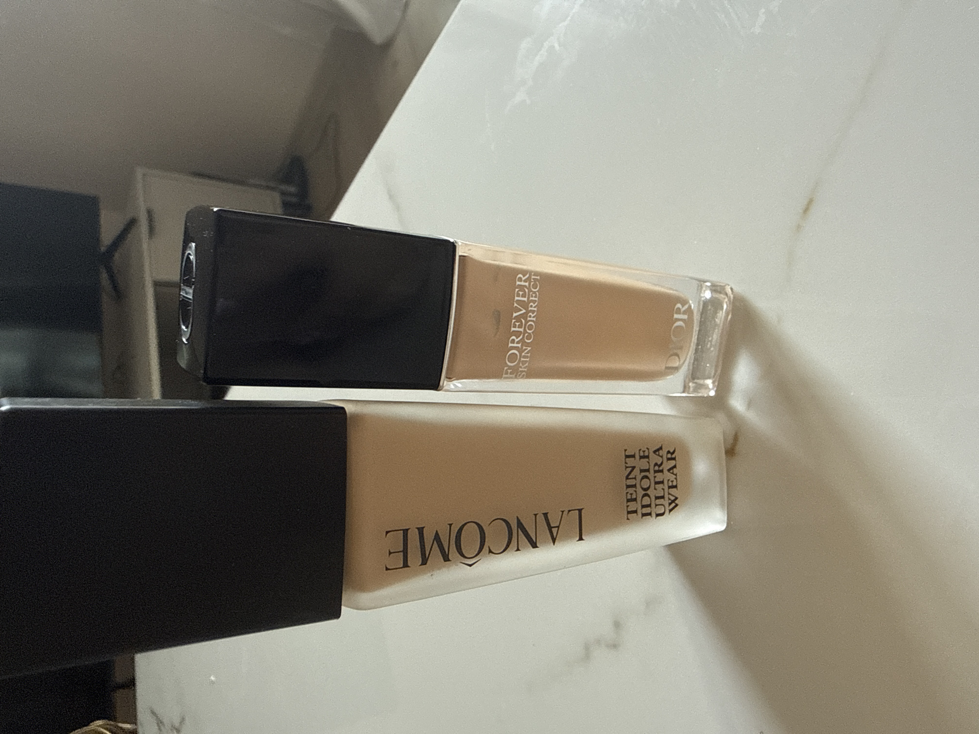 On an unidentified surface, possibly a countertop or table, are two bottles of makeup. The top bottle is Dior Forever Skin Correct, containing a light brown fluid. The bottom bottle is Lancome Teint Idole Ultra Wear, containing a light tan fluid. The location is in the city of Unknown, Unknown.