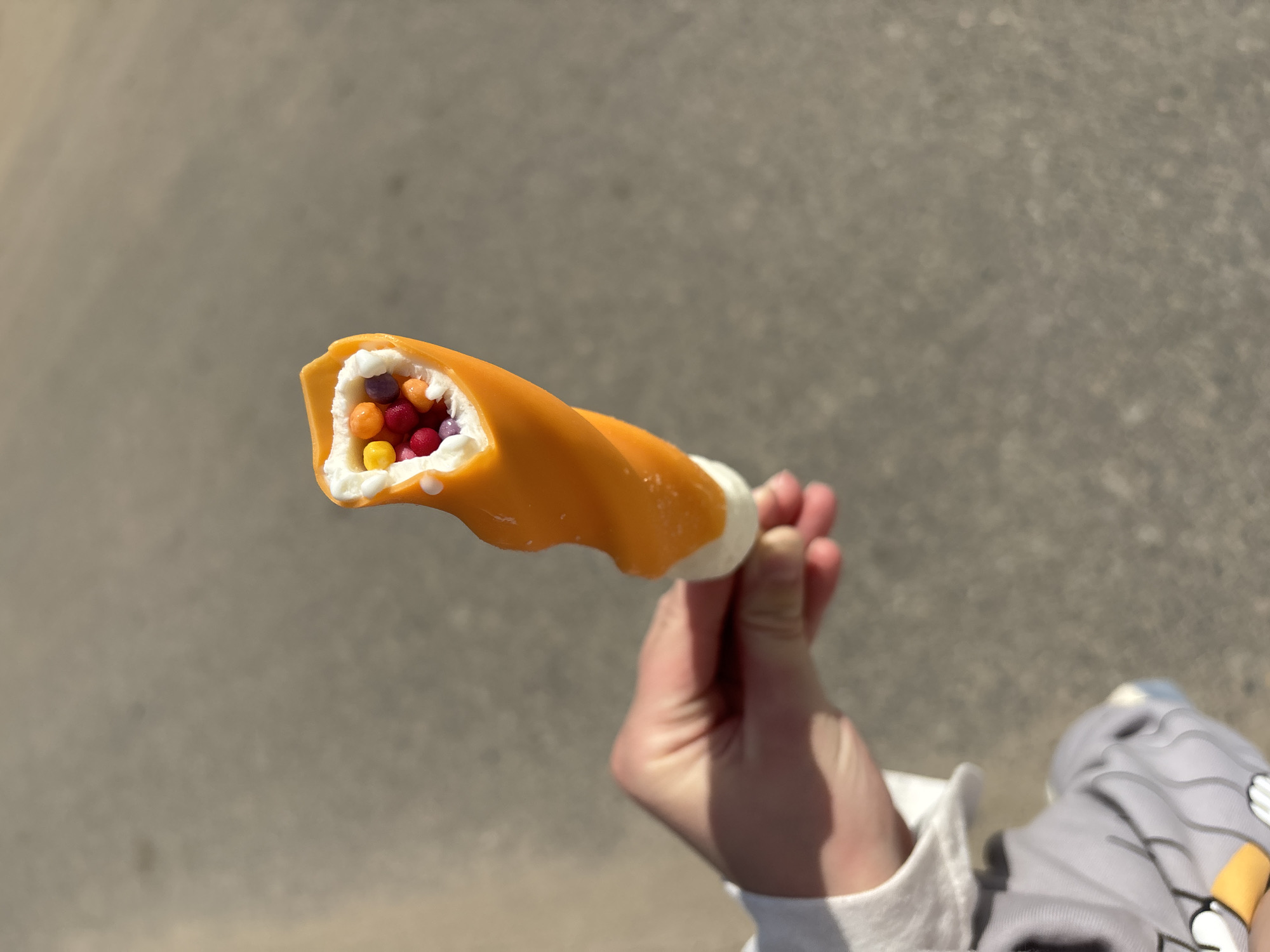 A hand holds an opened orange and white tube-shaped candy treat filled with small, multicolored, spherical candies. The background consists of a gray asphalt surface and a glimpse of clothing, suggesting the image was taken outdoors, possibly in a public area. There is no definitive information in the image to confirm the location is Шевлягино, Russia.