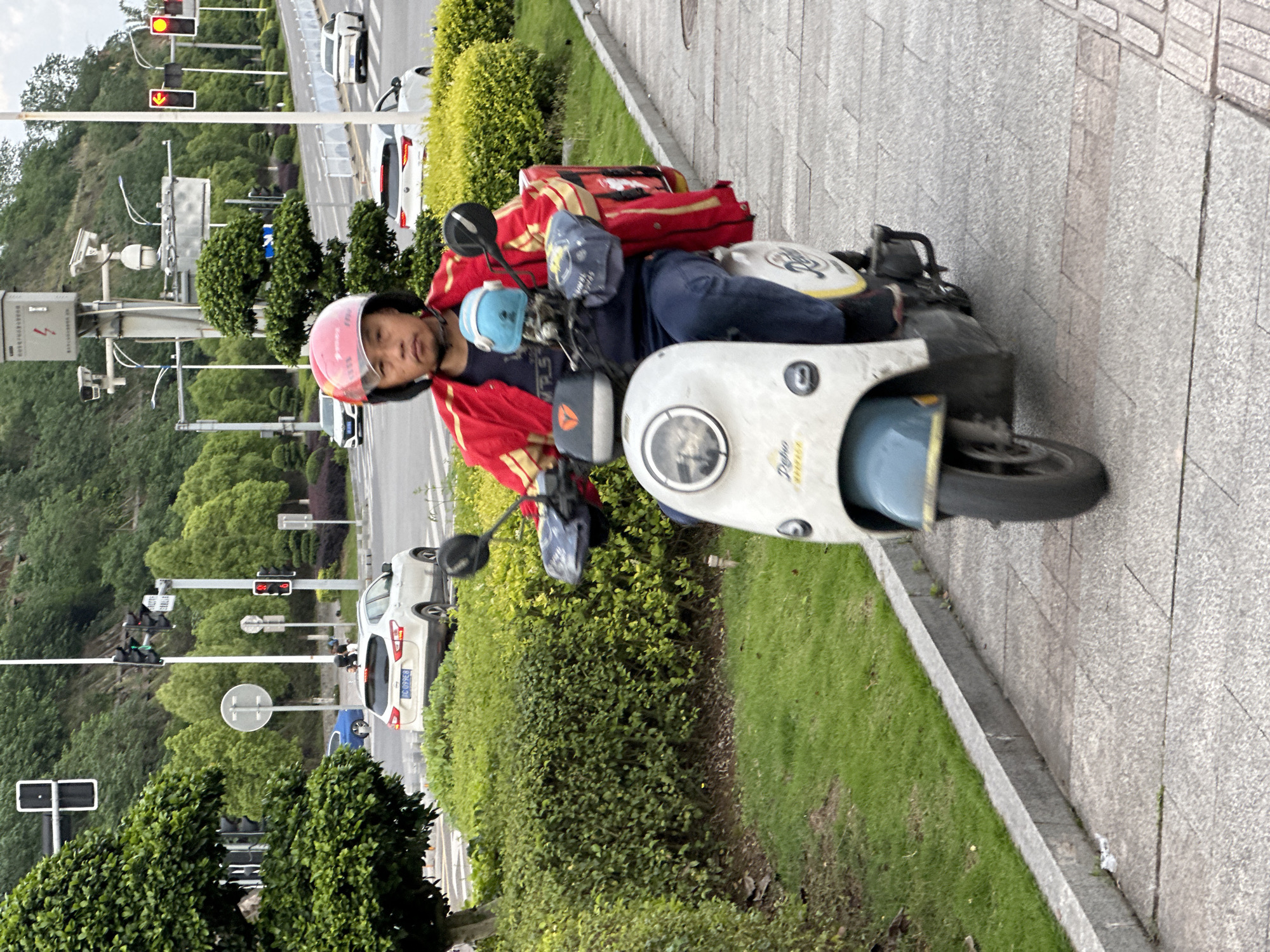 An outdoor eye-level shot captures a delivery driver on a scooter parked on a sidewalk next to a road, likely in Jiangjin, China. The driver, wearing a pink helmet, a red jacket, and blue pants, looks towards the camera with a neutral expression. A blue cap is visible on the scooter's handlebars. Behind the driver, a large red delivery bag with yellow accents is strapped to the back of the white and blue electric scooter. The scooter has "Deliro" and "ECHO" written on its front panel.
The scooter is positioned on a grey tiled sidewalk, adjacent to a strip of green grass and bushes. Beyond the bushes is a multi-lane road where several cars are stopped, suggesting a traffic light ahead. In the background, traffic lights show red signals, with one displaying a red downward arrow, indicating that traffic is halted. Various street poles, security cameras, and signs are visible along the roadside. One visible car has a license plate "BC 099ES". A blue road sign with a white right arrow is also discernible. Lush green trees cover a hillside in the far background. The scene appears to be taking place during the daytime, under an overcast sky, as indicated by the soft, diffused lighting and lack of strong shadows.