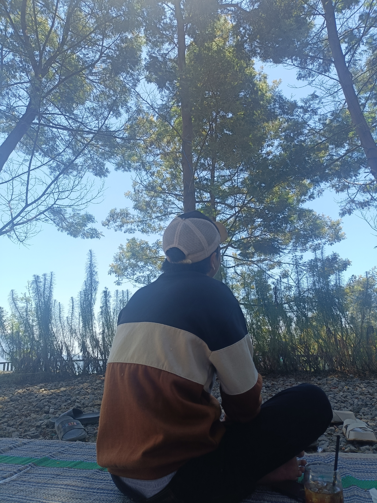 A person wearing a baseball cap and a color-blocked long-sleeved shirt sits cross-legged on a striped mat outdoors. They are facing away from the camera, looking up at tall trees against a clear blue sky. The ground around them is covered in small stones. Several pairs of sandals are visible on the stone surface. A glass with a dark beverage and ice, and a straw, sits on the mat near the person. The scene appears to be set in a natural, outdoor environment within the city of Kopeng, Indonesia, possibly a park or recreational area.
