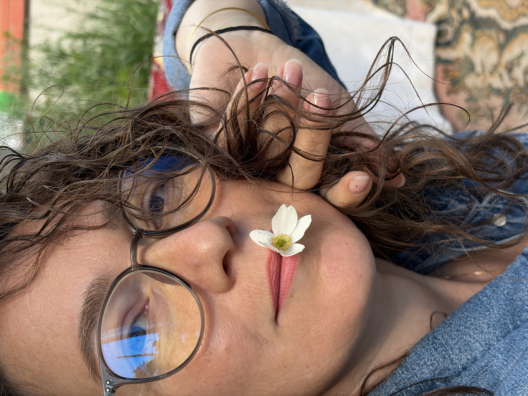 In Ol'khovskoye, Russia, a close-up, medium shot captures a woman in a denim jacket holding a white flower with a yellow center against her lips. Her brown, curly hair is partially covering her face, and she is wearing glasses with thick, round frames. The background is blurred, showing greenery and possibly a building. The woman's hand, with light-colored fingernails, is near her face, touching her hair. A thin gold bracelet is visible on her wrist. The lighting suggests an outdoor setting.
