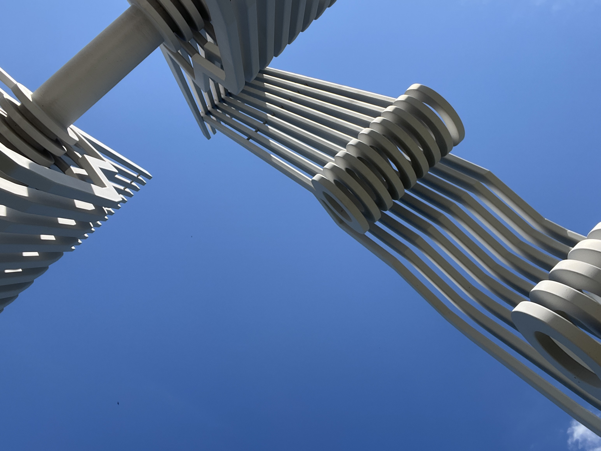 A low-angle, close-up shot of abstract, grey metal sculpture against a bright blue sky. The sculpture is comprised of repeating, curved horizontal elements that are layered and angled. The sky is clear except for a few wisps of white clouds at the bottom left corner. Two small dark specks, possibly birds, are visible in the sky. The sculpture appears to be the "Wings of the Future" monument located in Tolyatti, Russia, situated near the banks of the Samara River on Komsomolsky Highway.