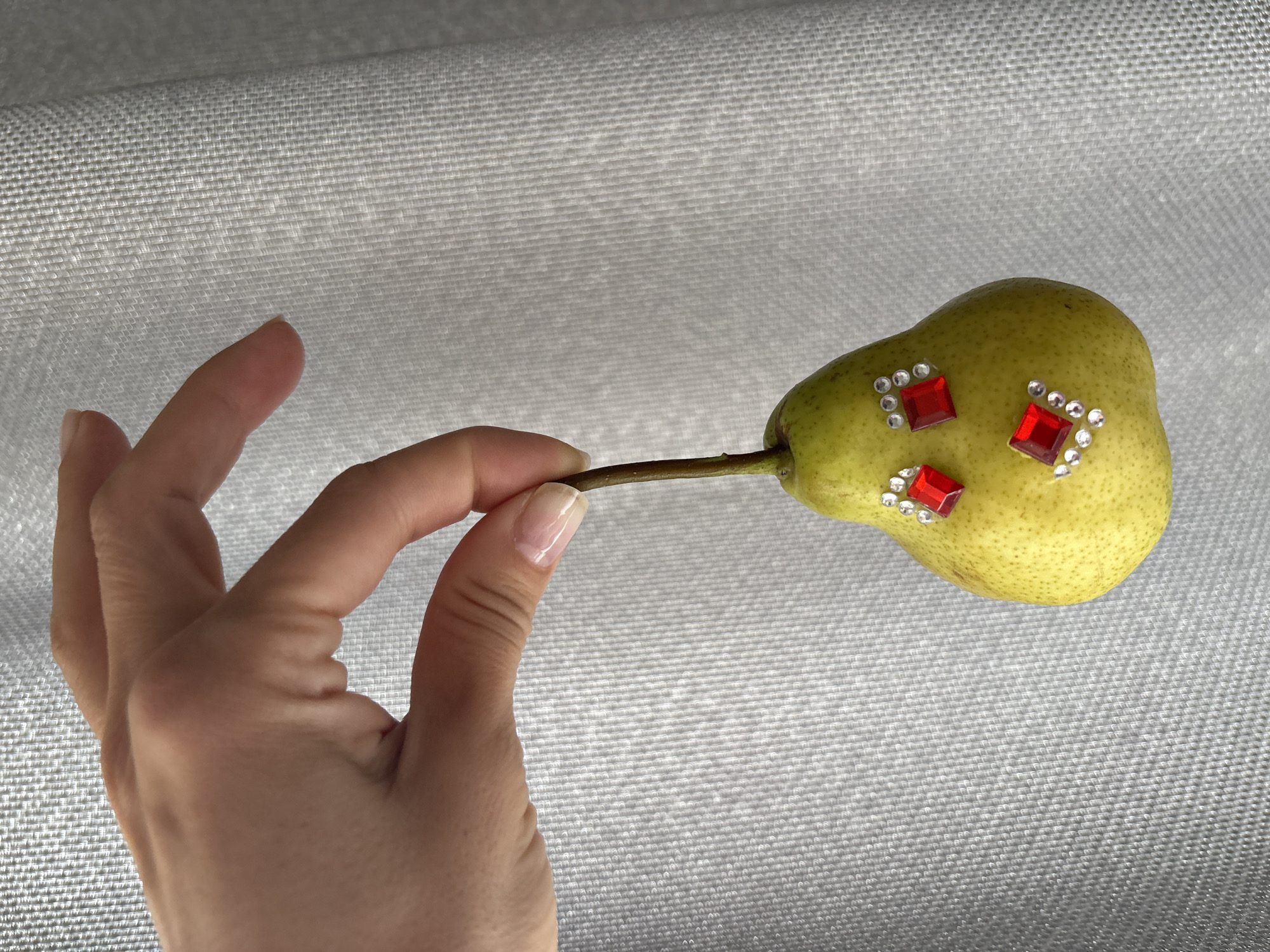 A close-up, eye-level shot, taken in Chaikovsky, Russia, features a hand holding a single, decorative pear against a textured, light-colored background.
The main subject is a greenish-yellow pear, held by its brown stem between the thumb and forefinger of a person's hand, positioned on the left side of the frame. The pear's skin shows natural speckles. It is strikingly embellished with three prominent red, square-shaped rhinestones, each surrounded by smaller, clear or white round rhinestones, creating a sparkling, artificial aesthetic on the natural fruit.
The hand, with natural-looking nails, is well-lit, revealing skin texture and subtle creases. The background is a uniform, light grey or off-white material, likely a woven fabric or embossed surface, displaying a fine, repeating grid or basketweave pattern. It is slightly out of focus, providing a soft contrast to the sharply rendered pear and hand. The lighting is soft and even, highlighting the details of the rhinestones and the pear's surface without harsh shadows. No specific time of day or outdoor weather is discernible, and there is no visible text in the image. The overall impression is that of an artistic or whimsical still life.