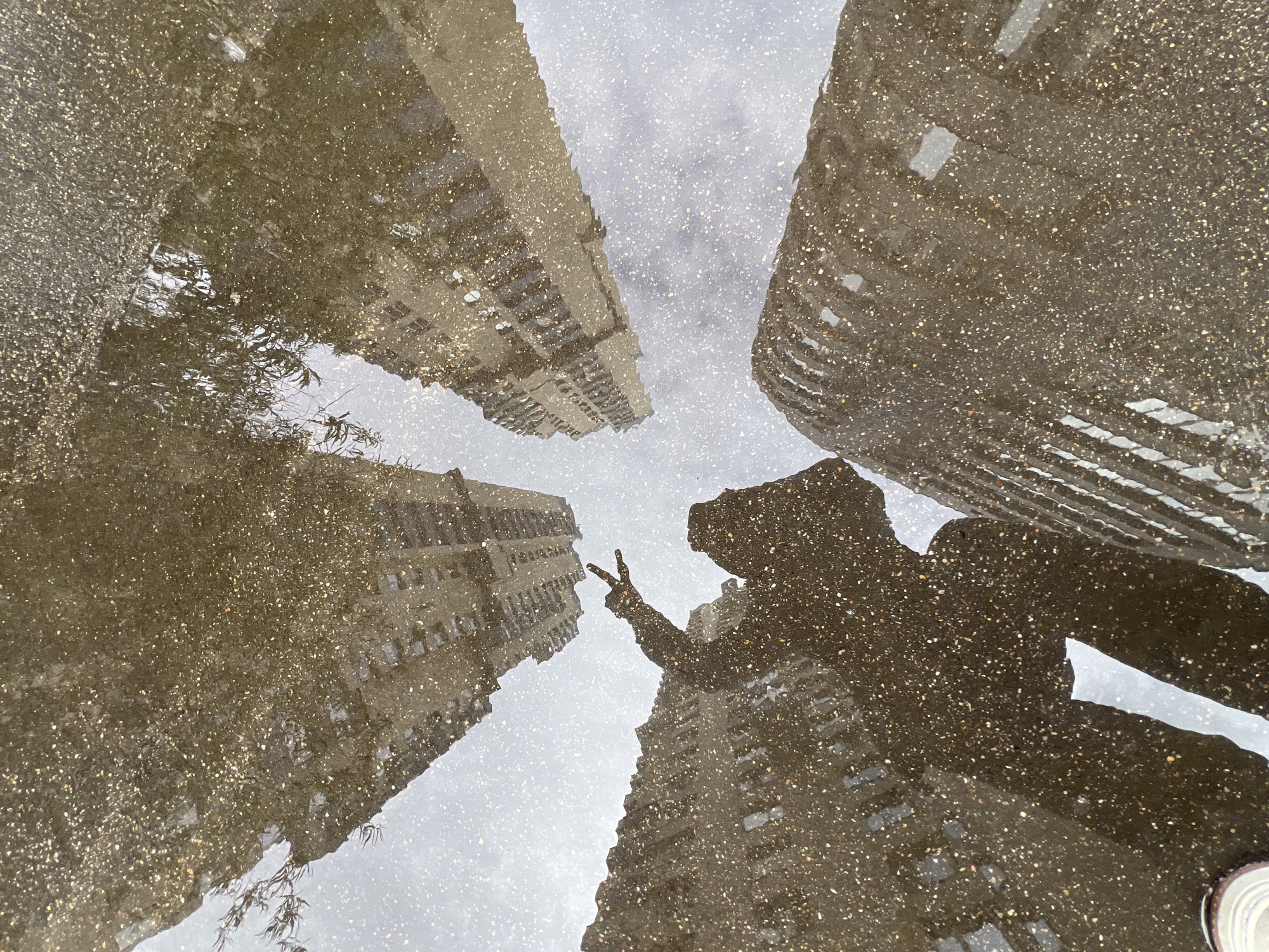The image shows a reflection in a puddle. The puddle reflects four buildings, three of which appear to be skyscrapers. A shadow of a person is visible, with their hand raised in a peace sign. The puddle is on a paved surface. The location is Moscow, Russia.