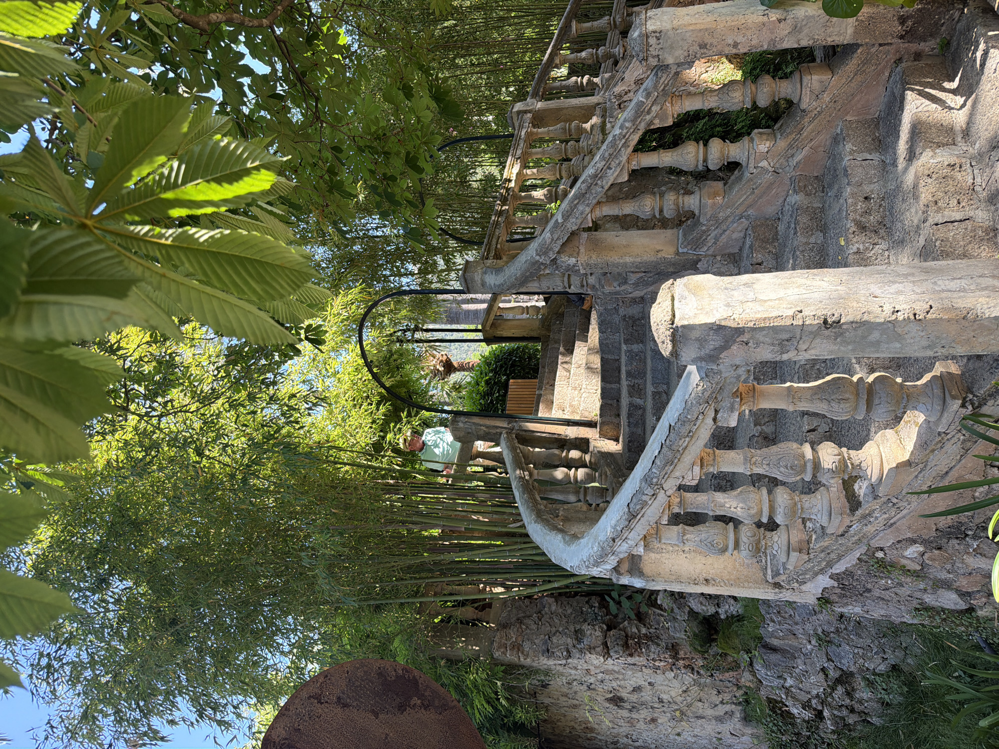 The image depicts a stone staircase with an ornate balustrade in a garden setting, likely in Bunyola, Spain. The stairs are aged and worn, leading upwards through lush vegetation that includes trees, bamboo, and other plants. Sunlight filters through the foliage, creating dappled patterns on the steps and balustrade. A person wearing a light-colored shirt can be seen partially obscured by the vegetation. In the foreground, a large, dark, possibly rusted object is partially visible. The scene conveys a sense of historical architecture integrated into a natural environment.